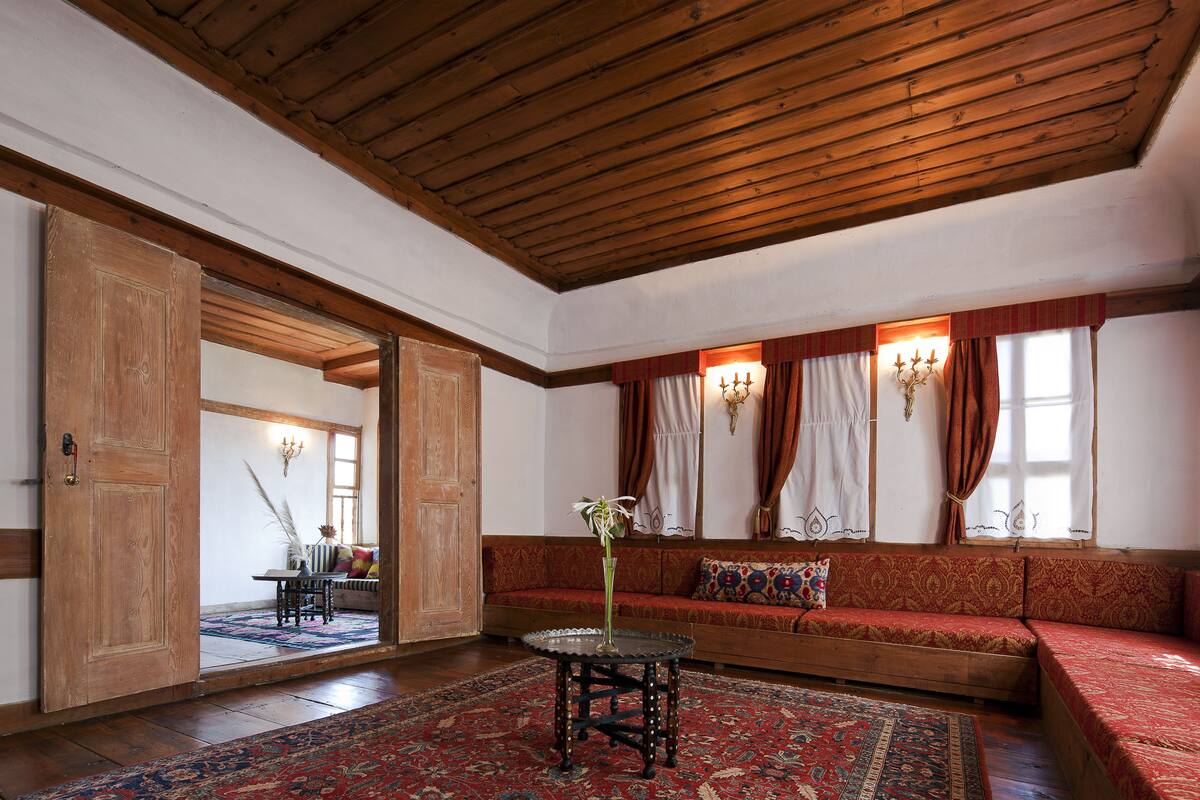 Una habitación con techo de madera, un sofá rojo con estampado, una mesa redonda con un jarrón y una ventana grande con cortinas.