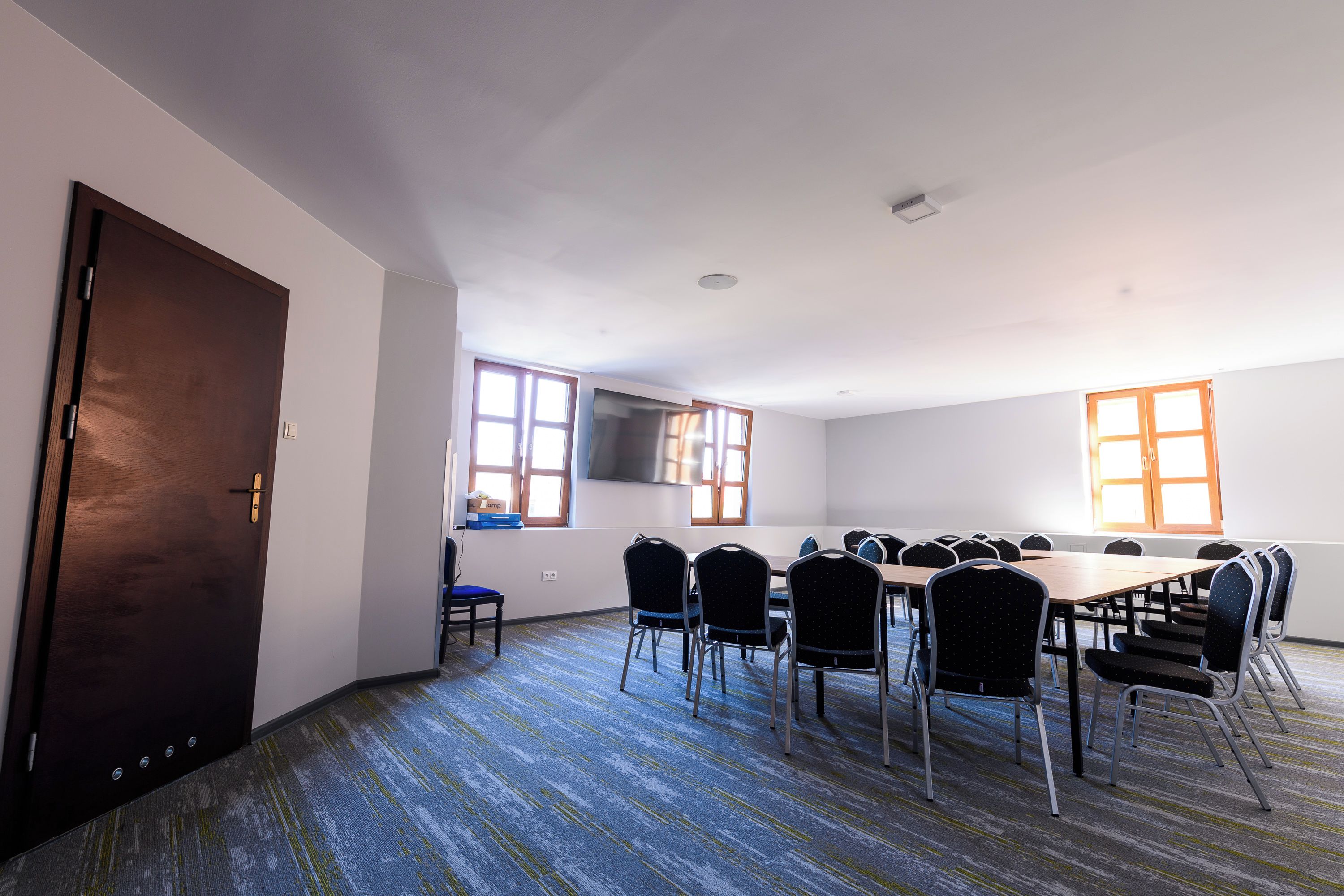A conference room with a large table and multiple chairs, a door, windows, and a flat-screen TV on the wall.