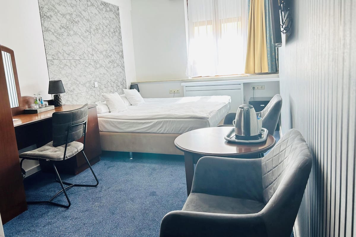 Quarto de hotel com cama, escrivaninha com luminária, cadeira, mesinha com chaleira e janela com cortinas.