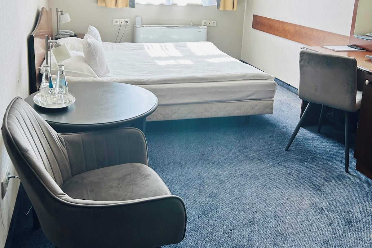 Quarto de hotel com cama, escrivaninha, cadeira e pequena mesa com garrafa e copos.