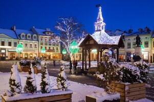 Una plaza cubierta de nieve con un pabellón, una aguja de iglesia y edificios al fondo.