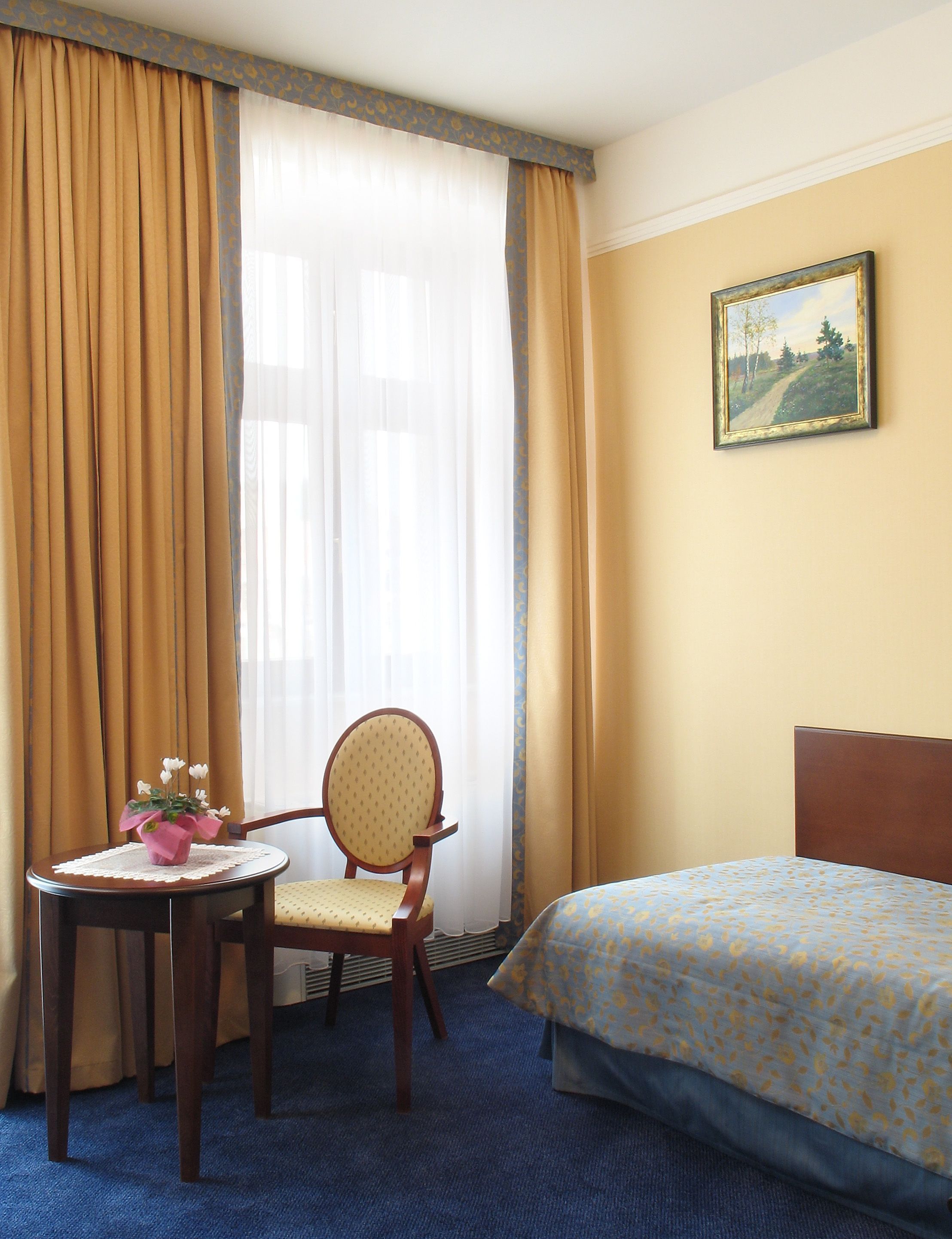 A hotel room with a bed, a chair, a small table with a vase of flowers, and a painting on the wall.