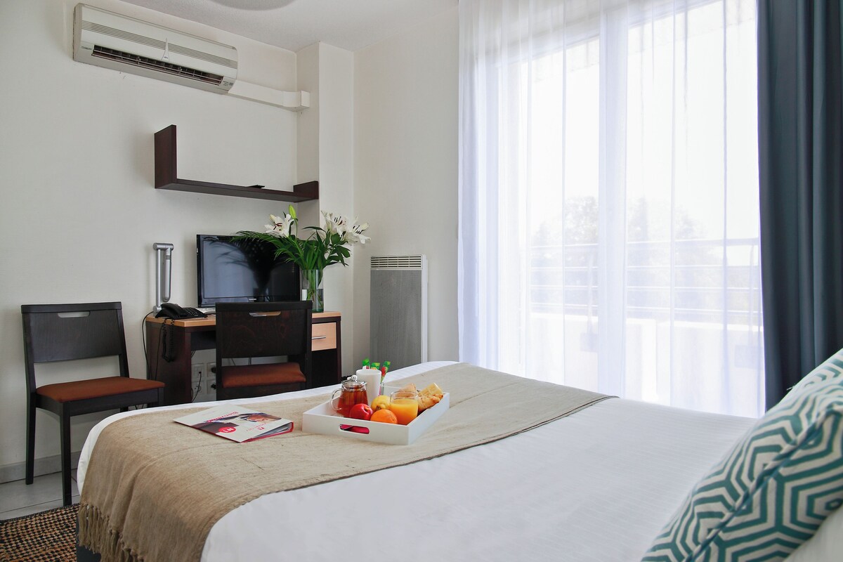 A hotel room with a bed, a desk with a computer, a chair, a bedside table with a phone, and a window with curtains.