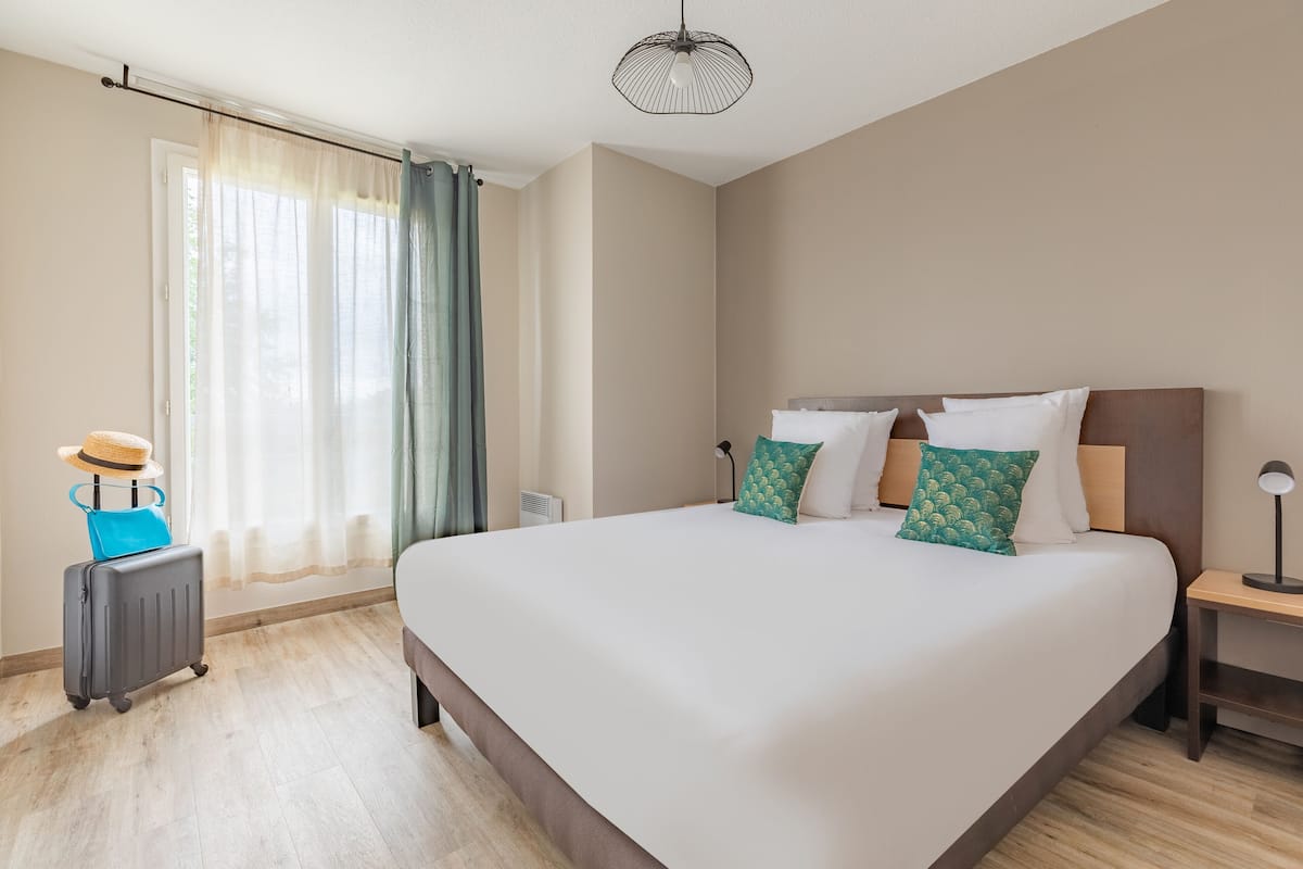 A large bed with a wooden headboard, a bedside table, and a suitcase with a hat on it.