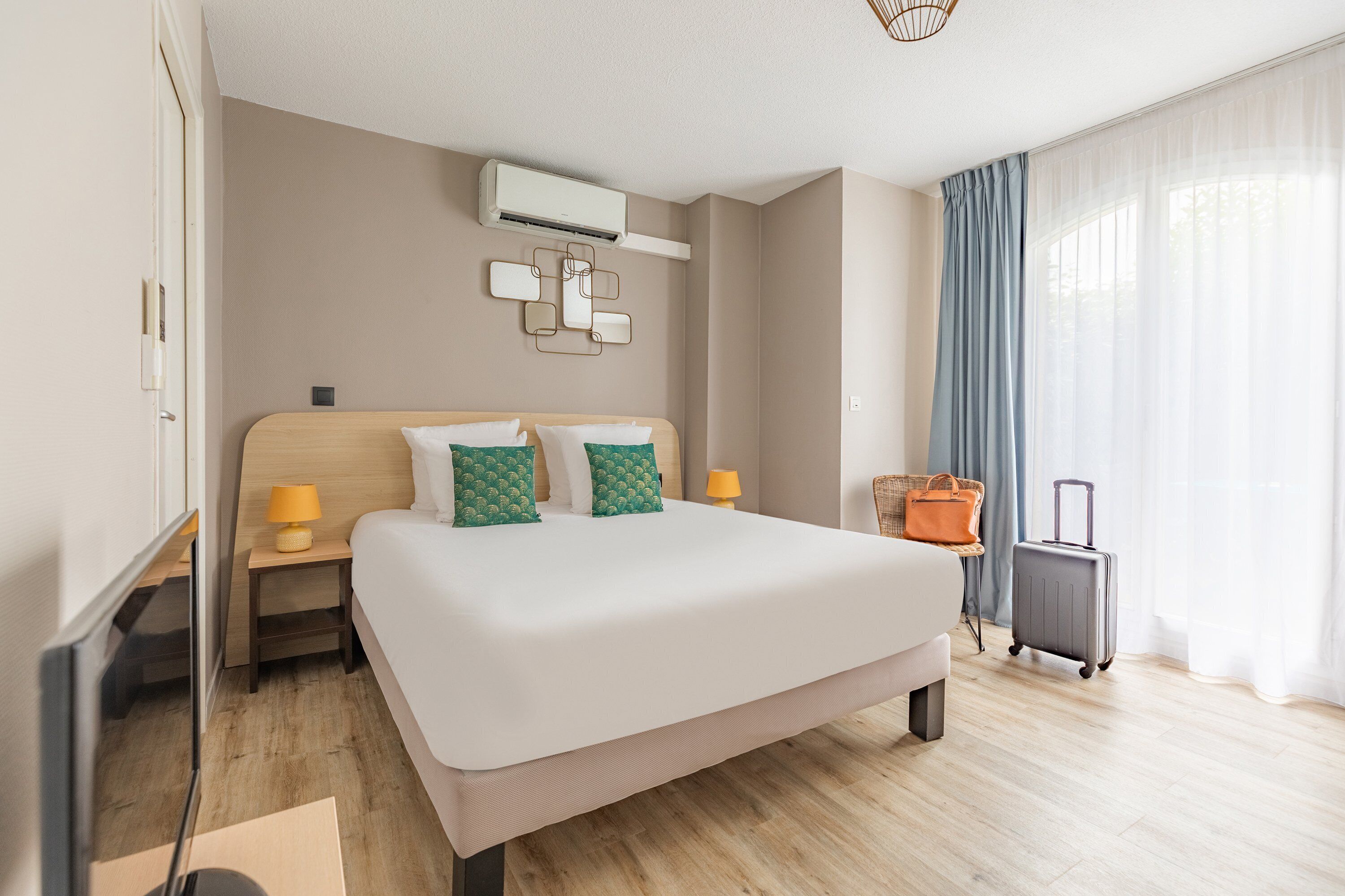A hotel room with a large bed, a flat-screen TV, a bedside table with a lamp, an air conditioning unit, and a suitcase.