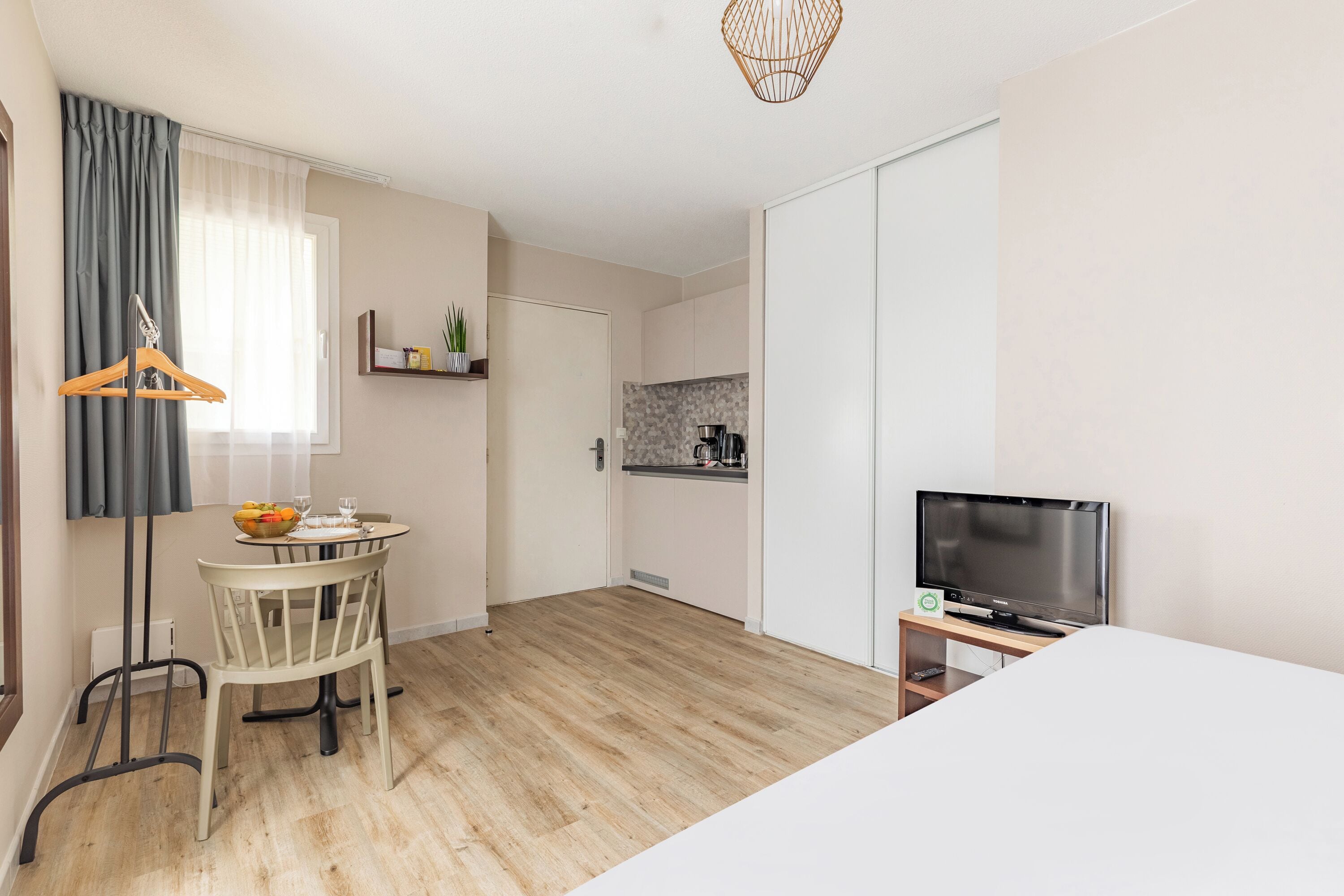 Ein modernes Hotelzimmer mit Holzboden, einem Essbereich mit Tisch und Stühlen, einem Fernseher auf einem Schrank und einer Küchenzeile mit Arbeitsfläche und Schränken.