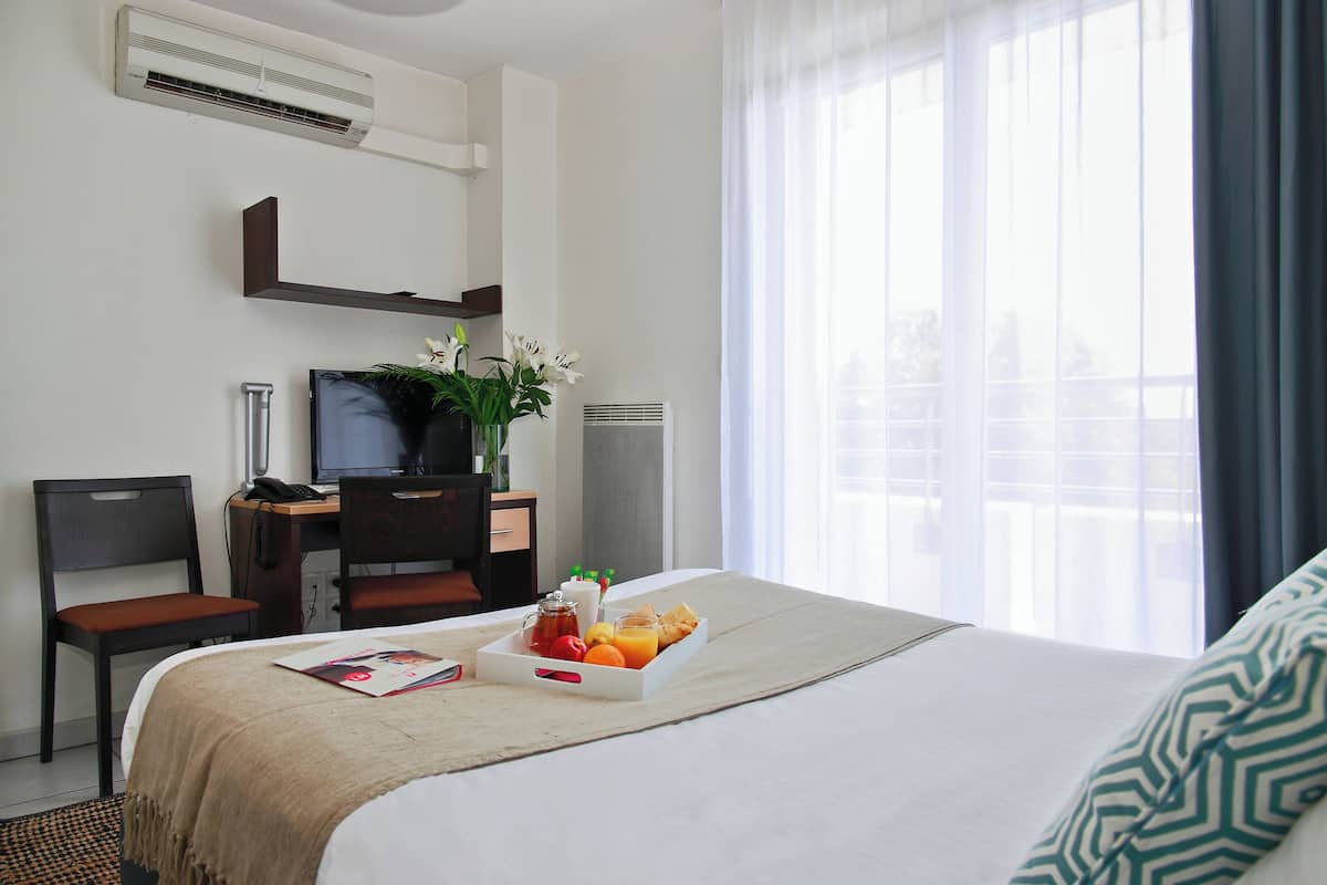 A hotel room with a bed, a desk with a computer, a chair, a bedside table with a phone, and a window with curtains.