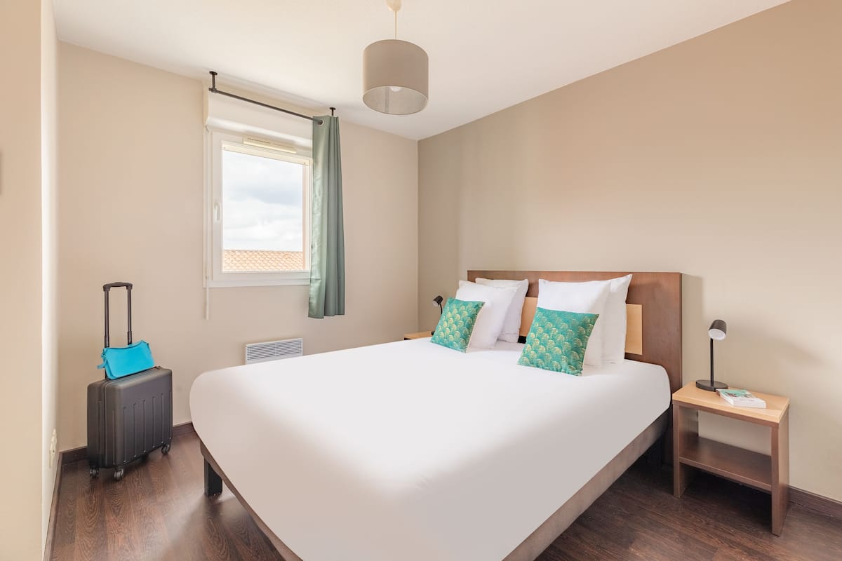 A hotel room with a large bed, a nightstand, a lamp, a suitcase, and a window with curtains.