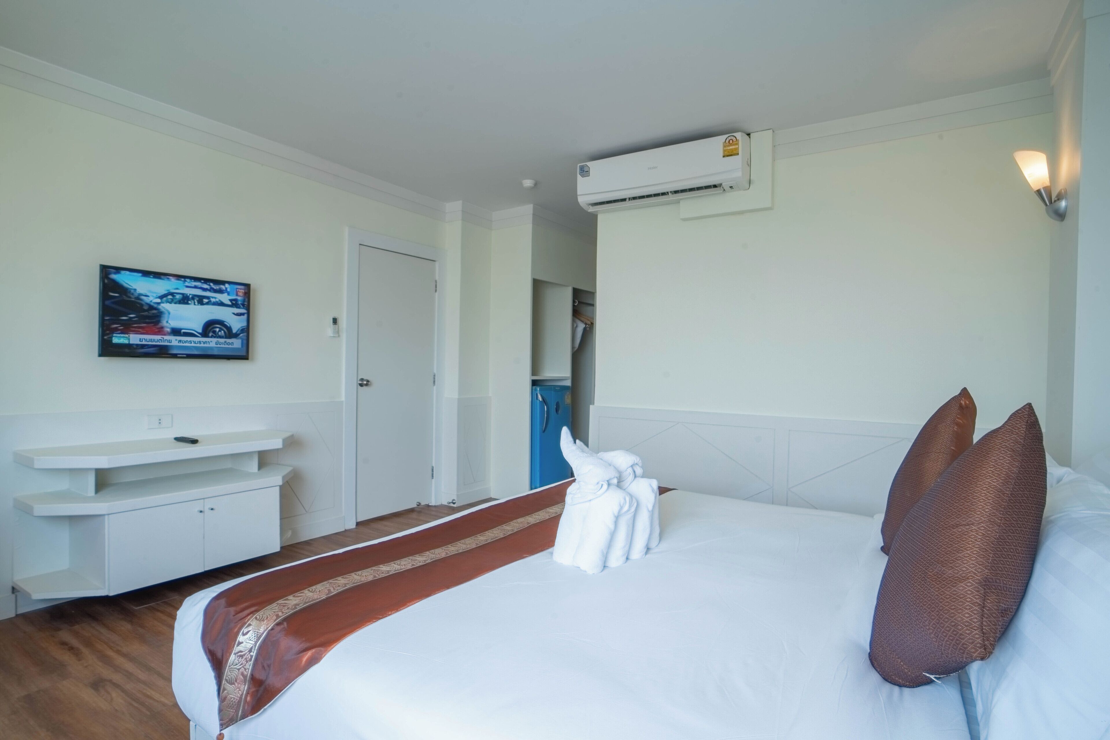 A hotel room with a bed, a TV mounted on the wall, a white console table, and an air conditioning unit.