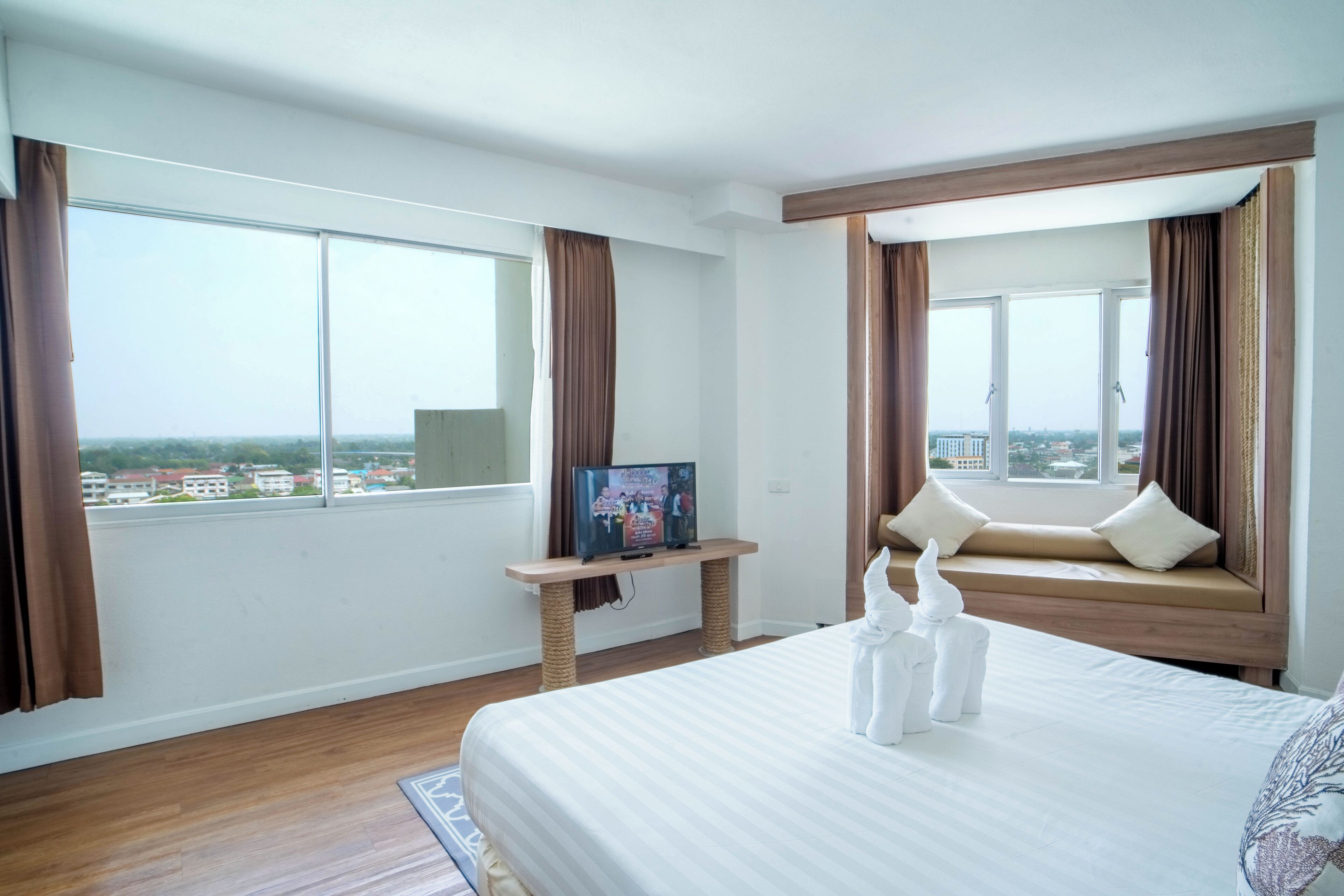 A hotel room with a large bed, a view of the city through the windows, a small table with a television, and a window seat with cushions.