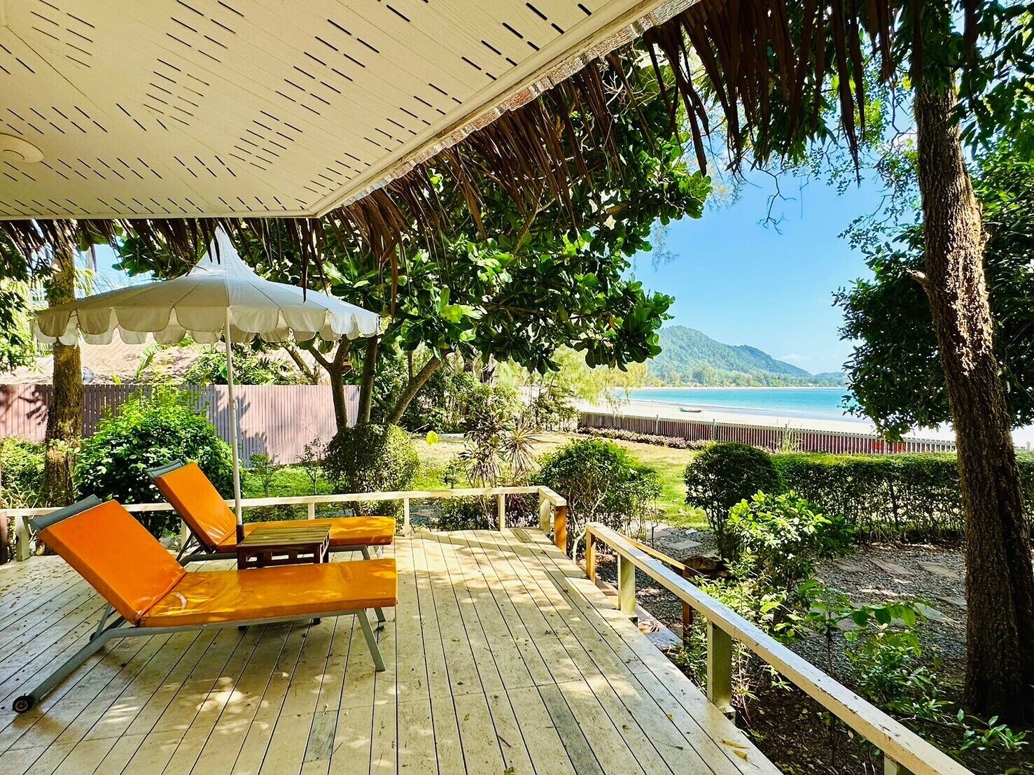 Una terraza de madera con dos sillones naranja y un paraguas blanco. Un cerco de madera y vegetación rodean la terraza. Al fondo se ve una playa con vista clara al océano y montañas distantes.