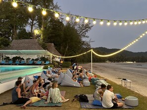 A poolside area with lounge chairs and bean bags, string lights overhead, and a view of the beach and hills in the background.