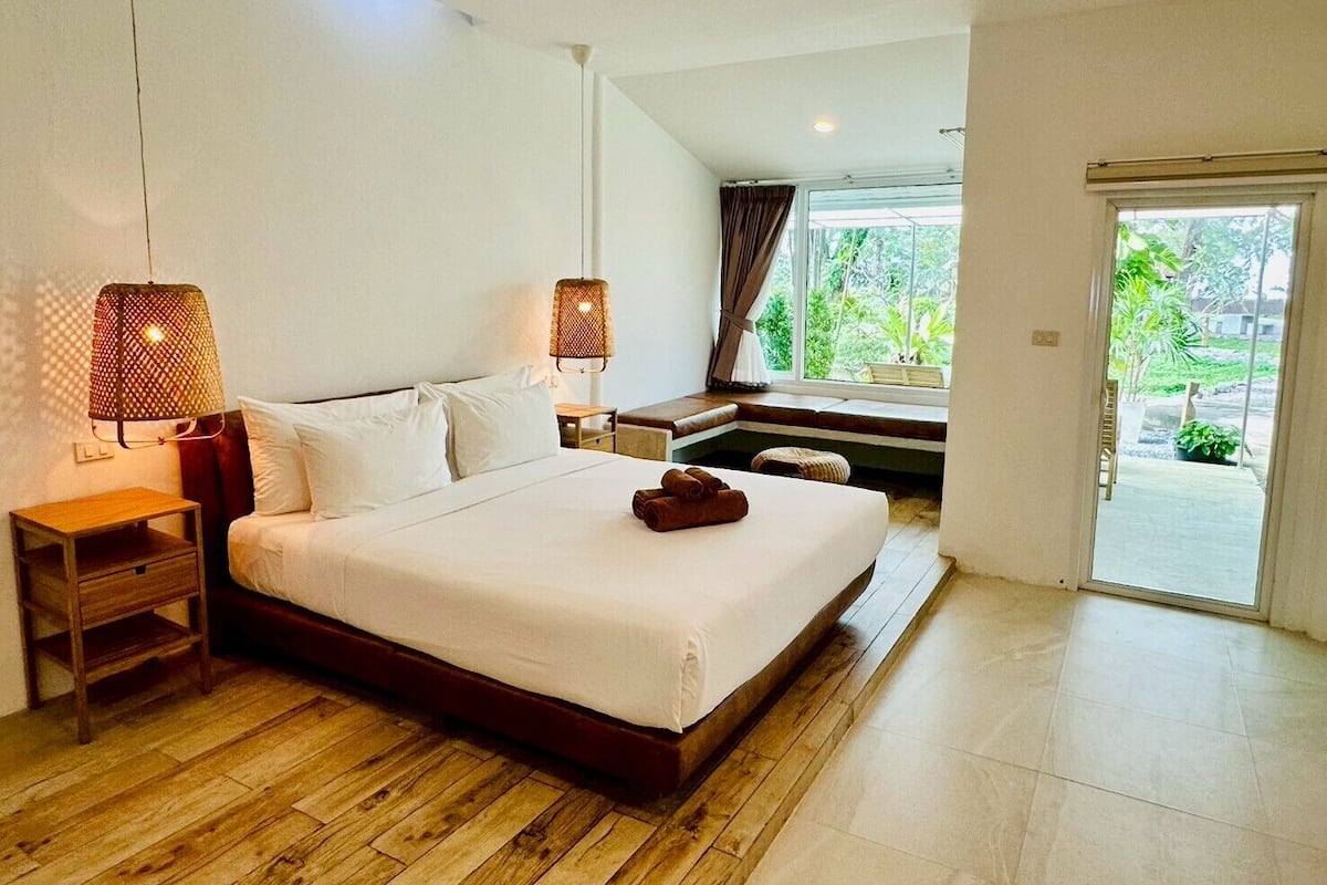 A bedroom with a large bed, two bedside tables, a window with curtains, and a view of greenery outside.