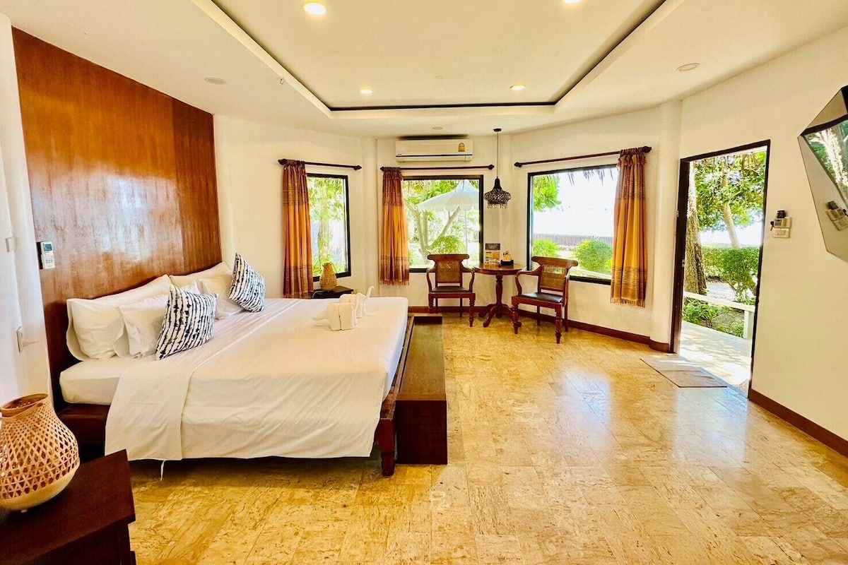 A hotel room with a large bed, a small table with chairs, and a view of greenery through the windows.
