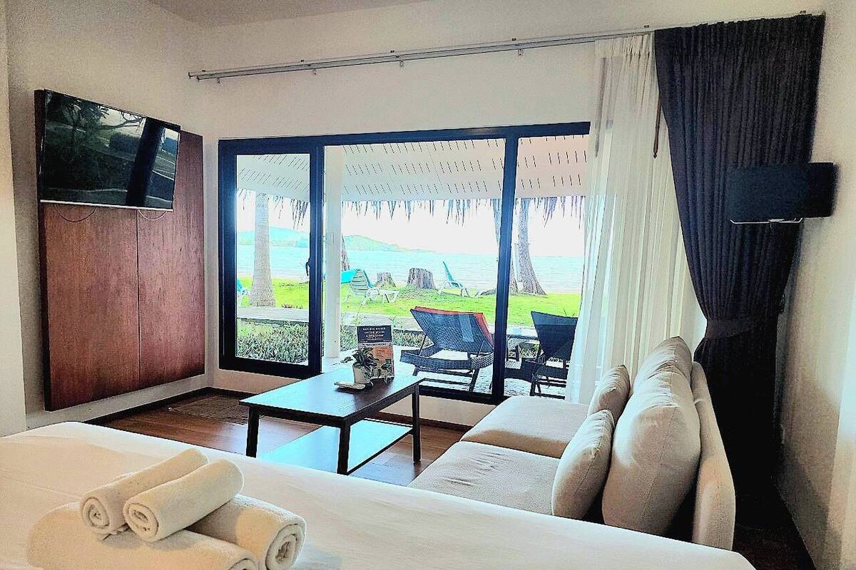 A hotel room with a large bed, a TV, a table, and a view of the sea through large windows.