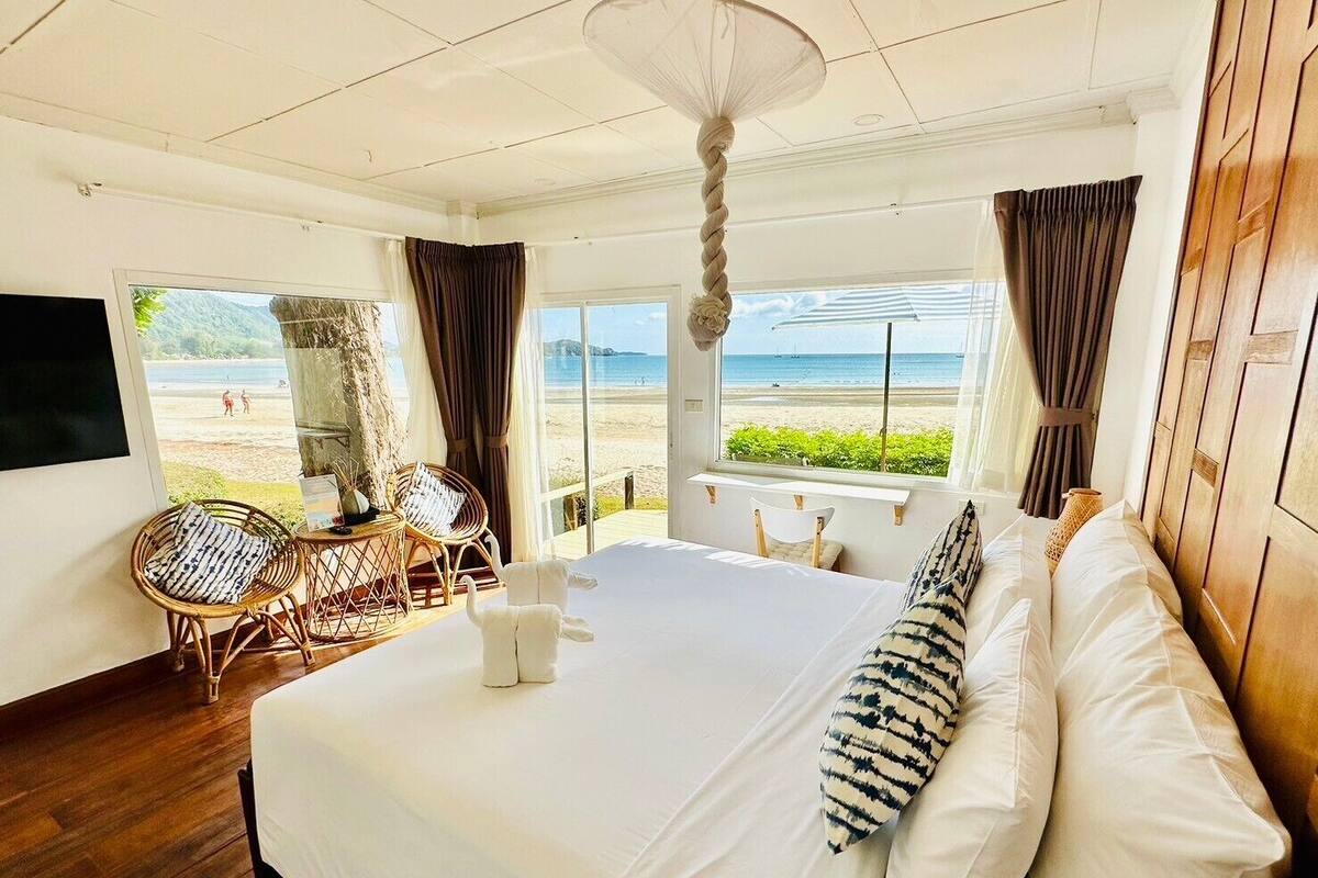 A hotel room with a large bed, a view of the beach, wicker furniture, and a ceiling fan.