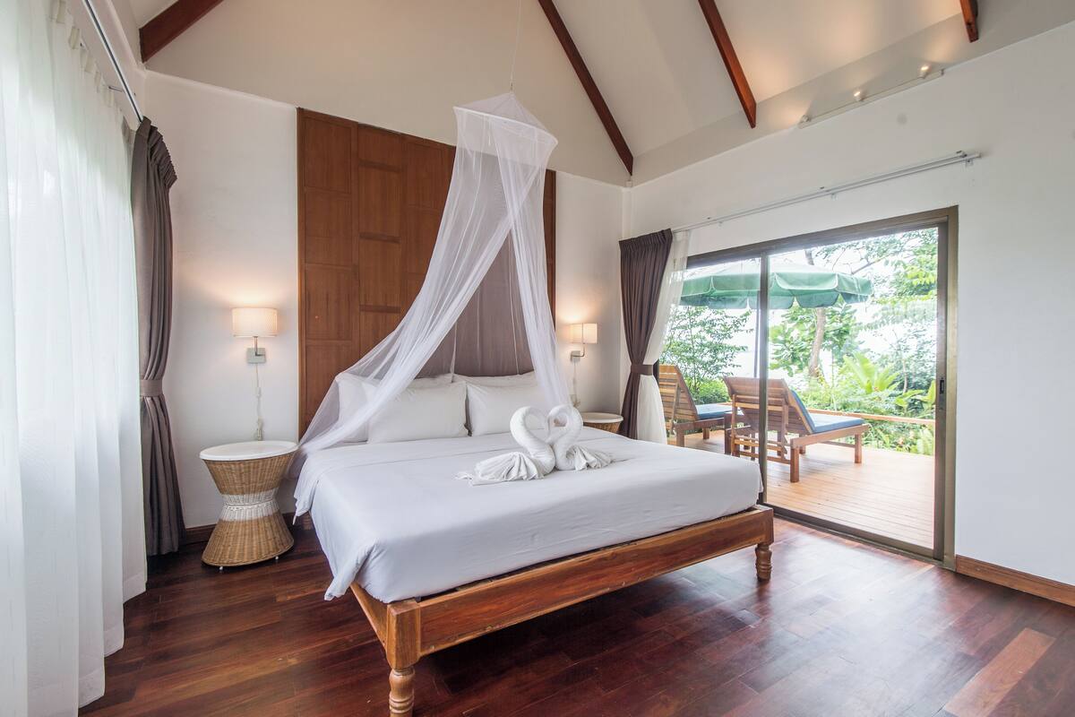 A large bed with a canopy, a wicker side table, and a view of an outdoor deck with chairs.
