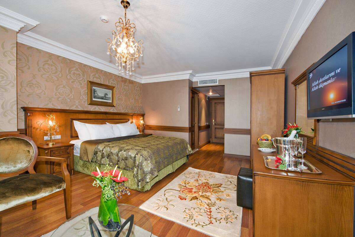Una habitación de hotel con cama, mesita de noche de madera, silla, escritorio con televisor y un jarrón decorativo.
