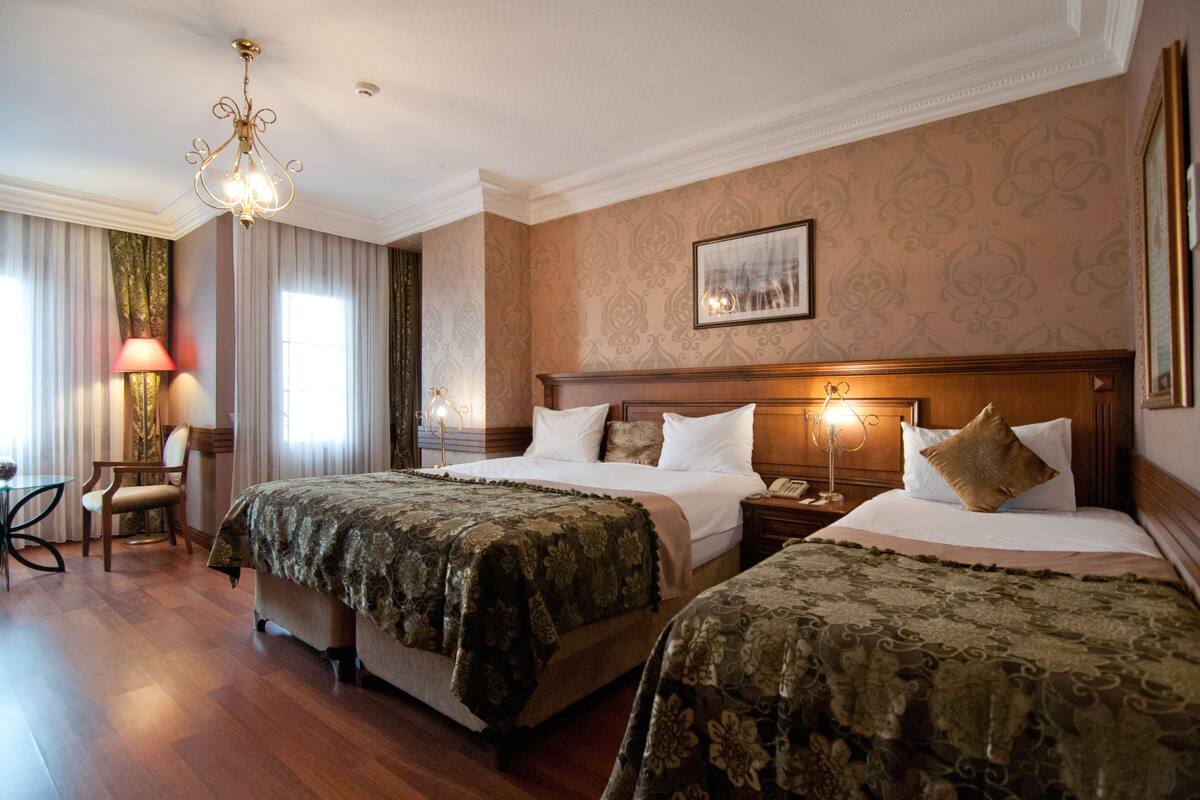 Habitación de hotel con dos camas, cabecera de madera, candelabro, escritorio con silla y ventana con cortinas.
