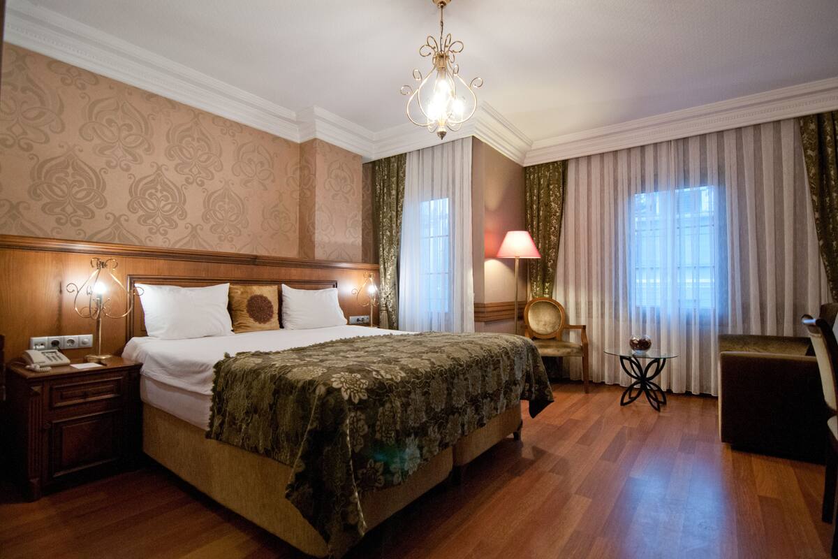 Una habitación de hotel con una cama grande, una mesita de noche, una lámpara, una silla, una pequeña mesa y una araña.