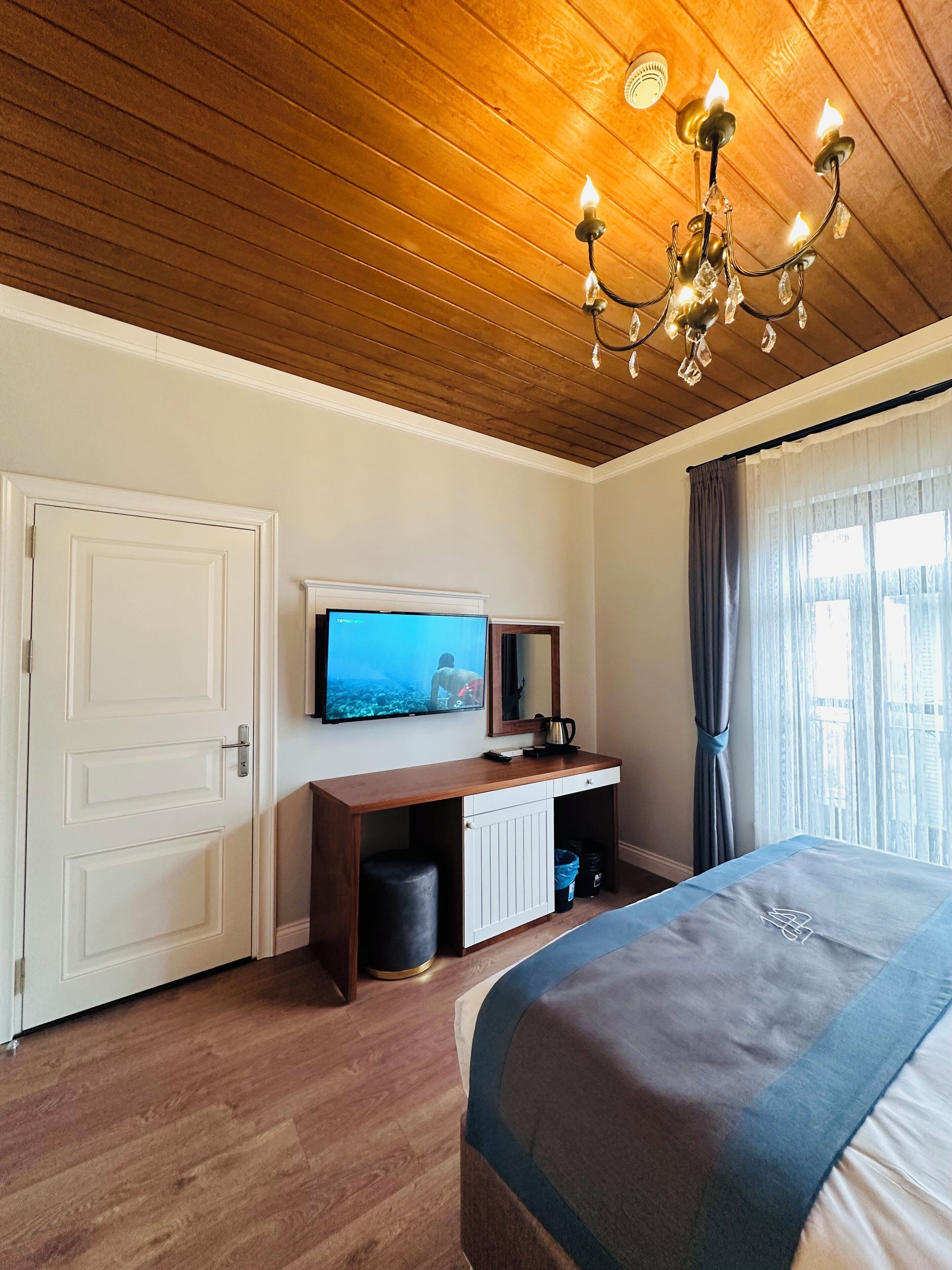 Habitación de hotel con techo de madera, una cama, un televisor sobre un mueble blanco, una ventana con cortinas y una puerta.