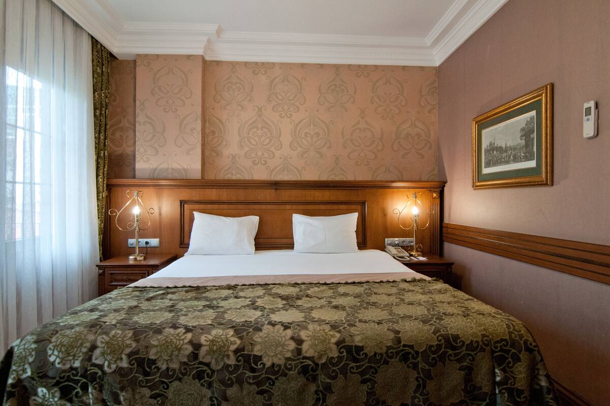 Habitación de hotel con una cama grande, dos lámparas de noche, un cuadro enmarcado en la pared y una ventana con cortinas.