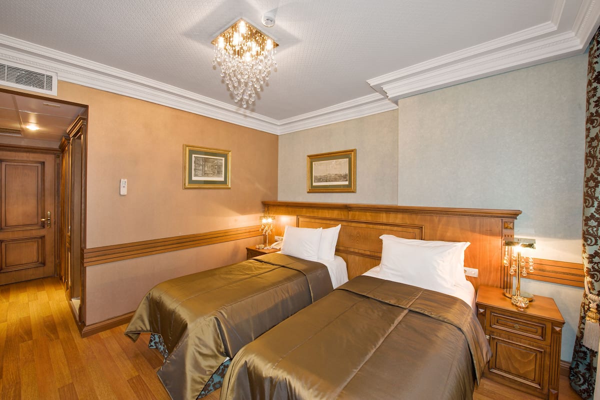 Habitación de hotel con dos camas, una araña, cuadros enmarcados en las paredes y una puerta de madera.