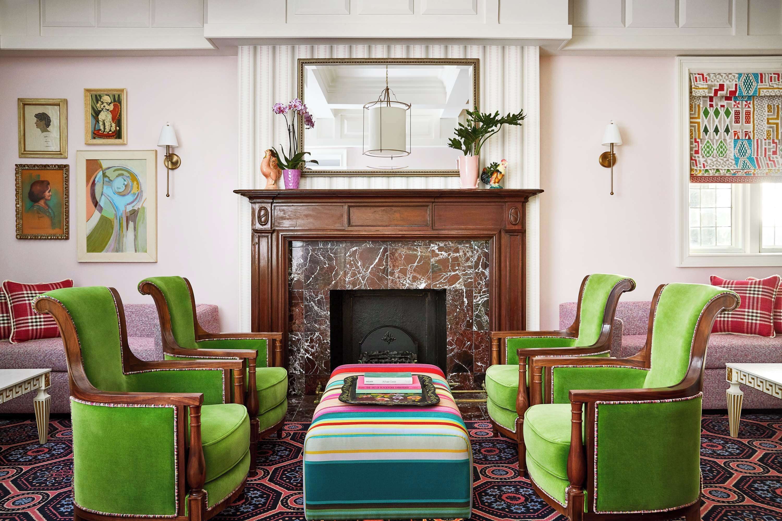 A room with a fireplace, green upholstered chairs, a striped ottoman, and artwork on the walls.
