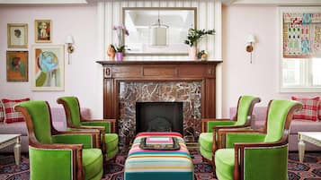 A room with a fireplace, green upholstered chairs, a striped ottoman, and artwork on the walls.