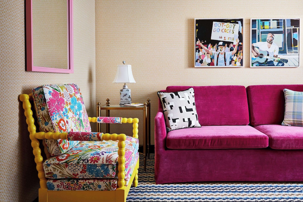 A living room with a vibrant floral armchair, a pink velvet sofa, a small side table with a lamp, and two framed artworks on the wall.