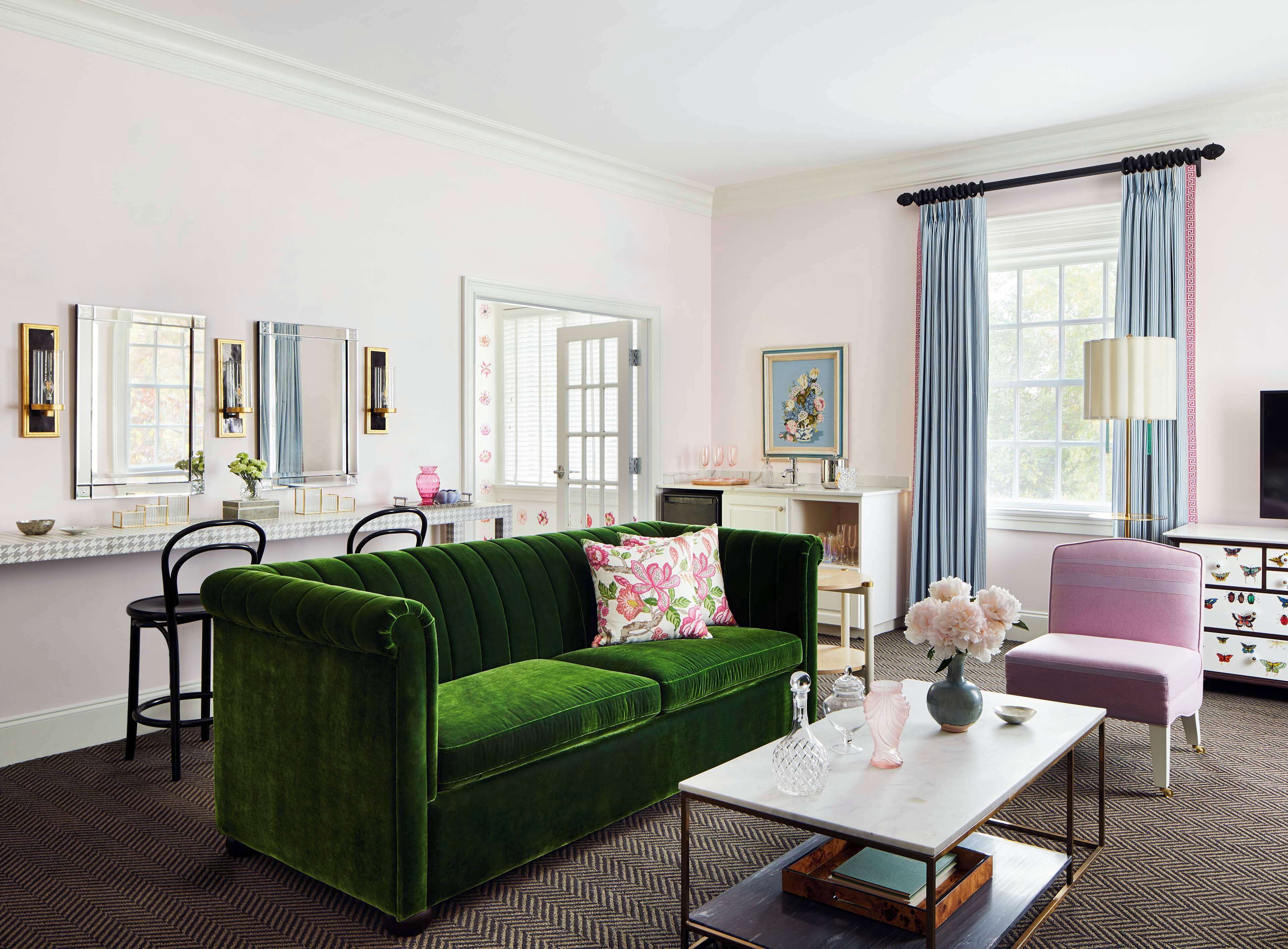Una habitación bien iluminada con un sofá verde, una pequeña mesa y una silla rosa. Se ve una ventana con cortinas, un espejo y un televisor.