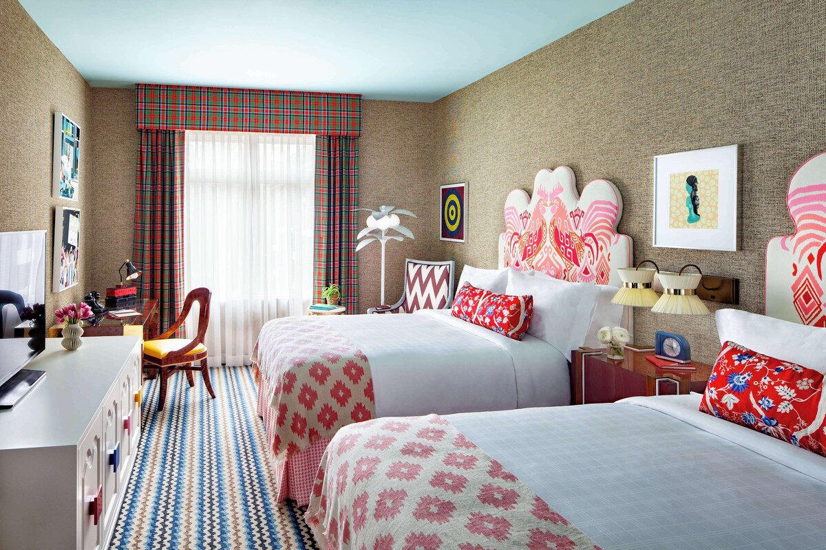 A hotel room with two beds, a desk, a chair, and a window with plaid curtains.