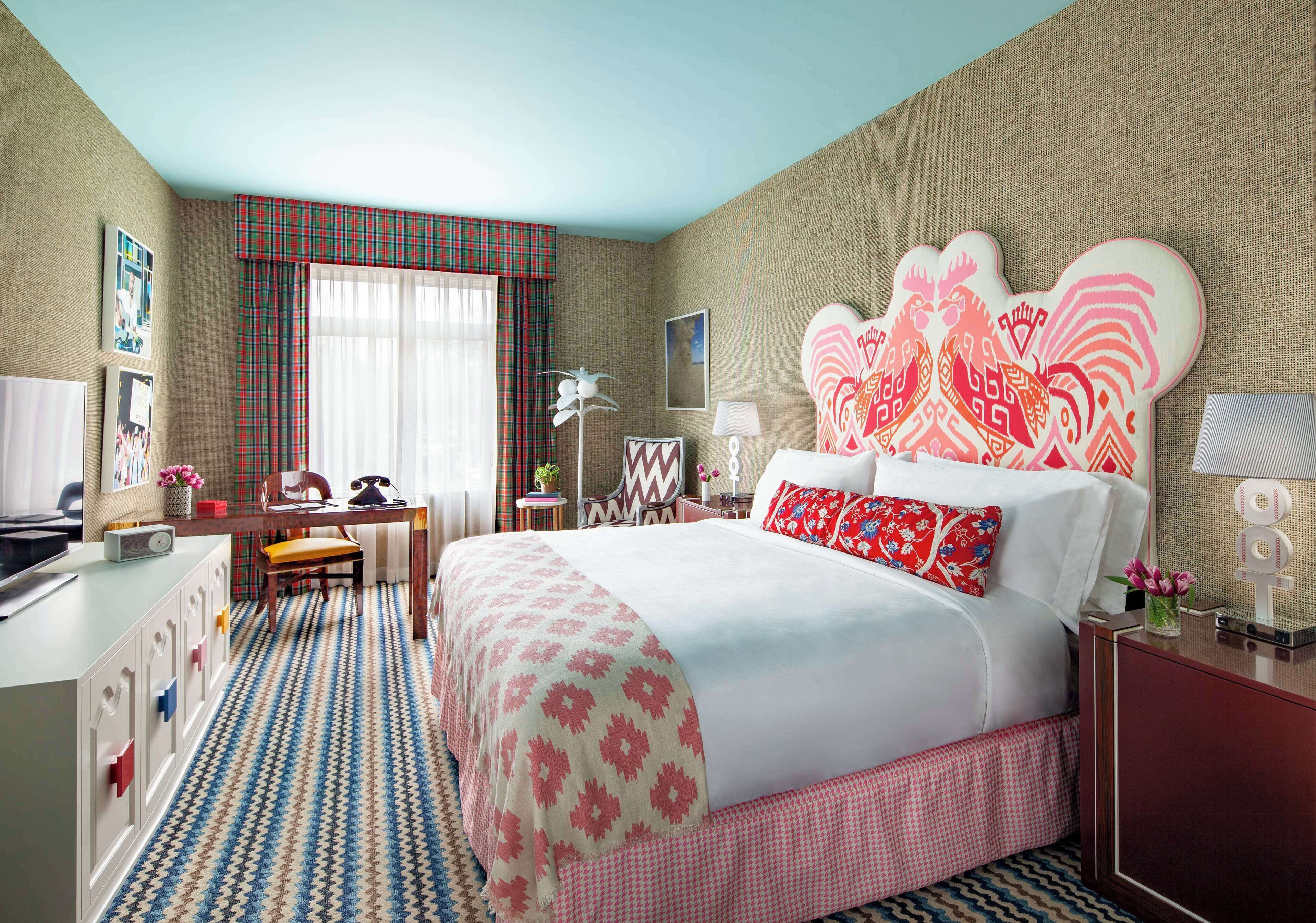 Una habitación de hotel con cama, escritorio, silla, lámpara de escritorio y ventana con cortinas.