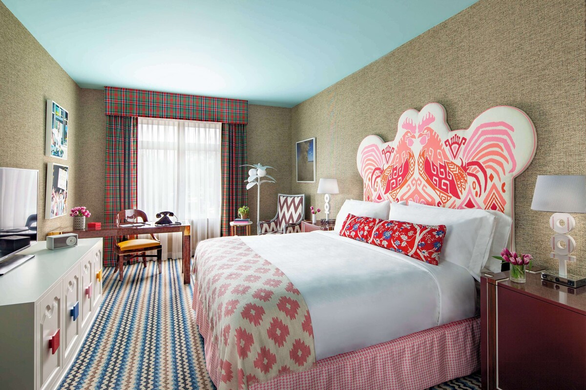 A hotel room with a bed, a desk, a chair, a desk lamp, and a window with curtains.