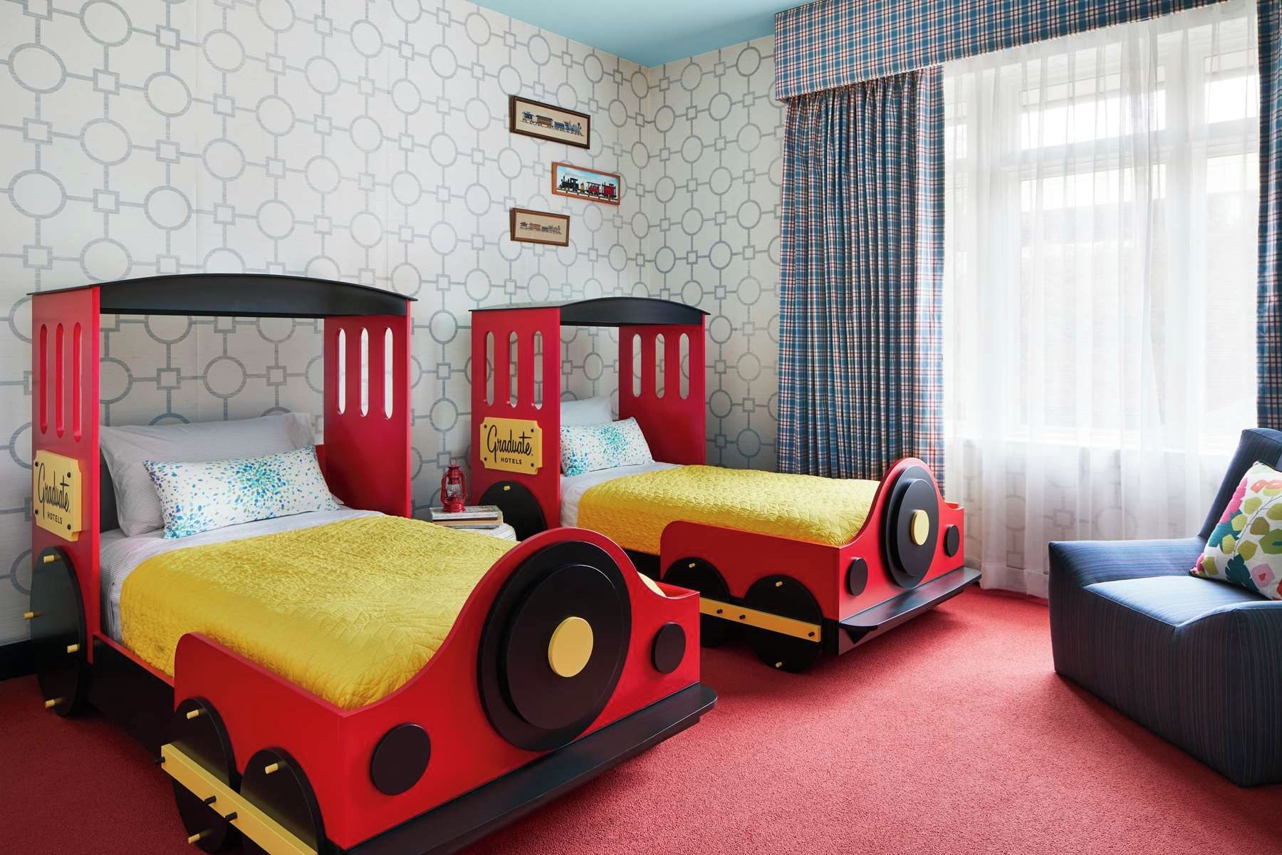 Habitación con dos camas diseñadas como trenes, alfombra roja y una ventana con cortinas.