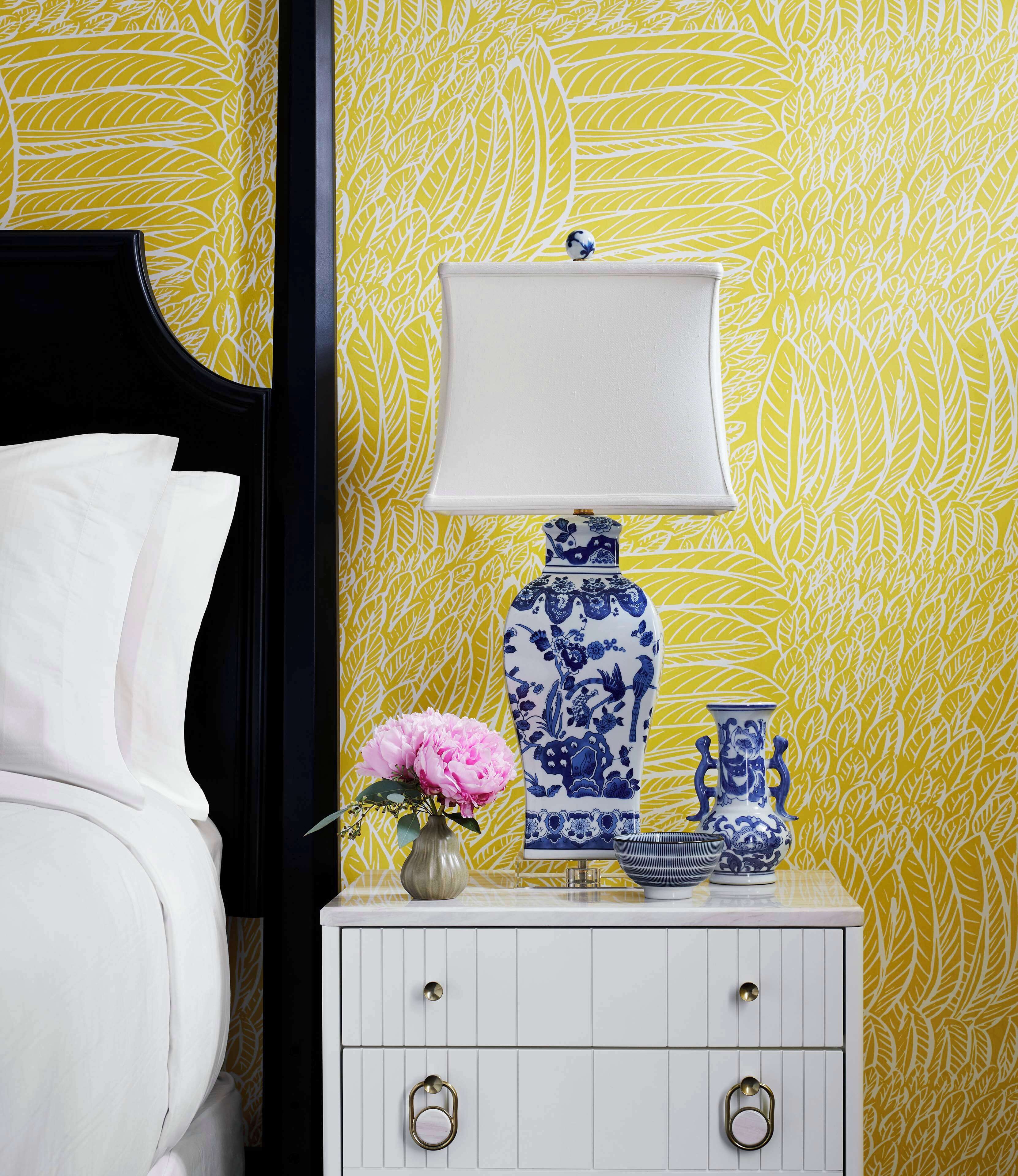 Une table de chevet blanche, ornée d’un vase bleu et blanc, d’une fleur rose et d’une lampe blanche, le tout placé devant un mur jaune à motifs de feuilles.