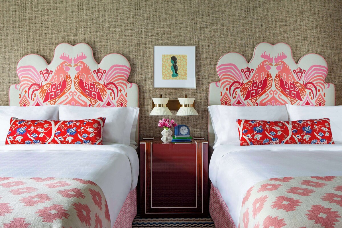 A hotel room with two beds, each with a unique patterned headboard, a nightstand with a clock and a vase of flowers, and a textured wall.