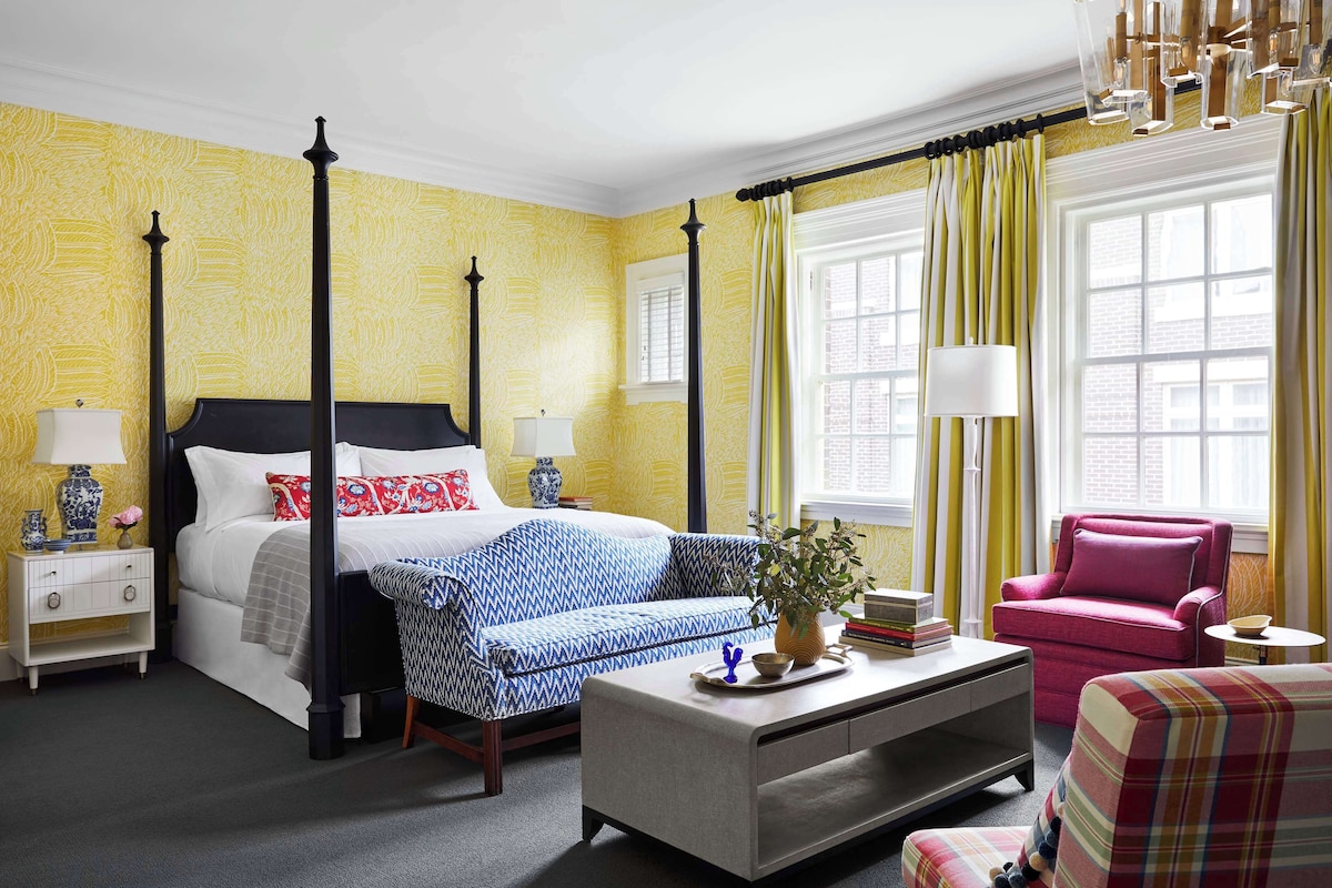 A four-poster bed with a blue and white patterned bedspread, a bedside table with a lamp, a small bedside table with a vase, a sofa with a blue and white patterned throw, a coffee table with a vase and books, a chair, and a window with yellow curtains.