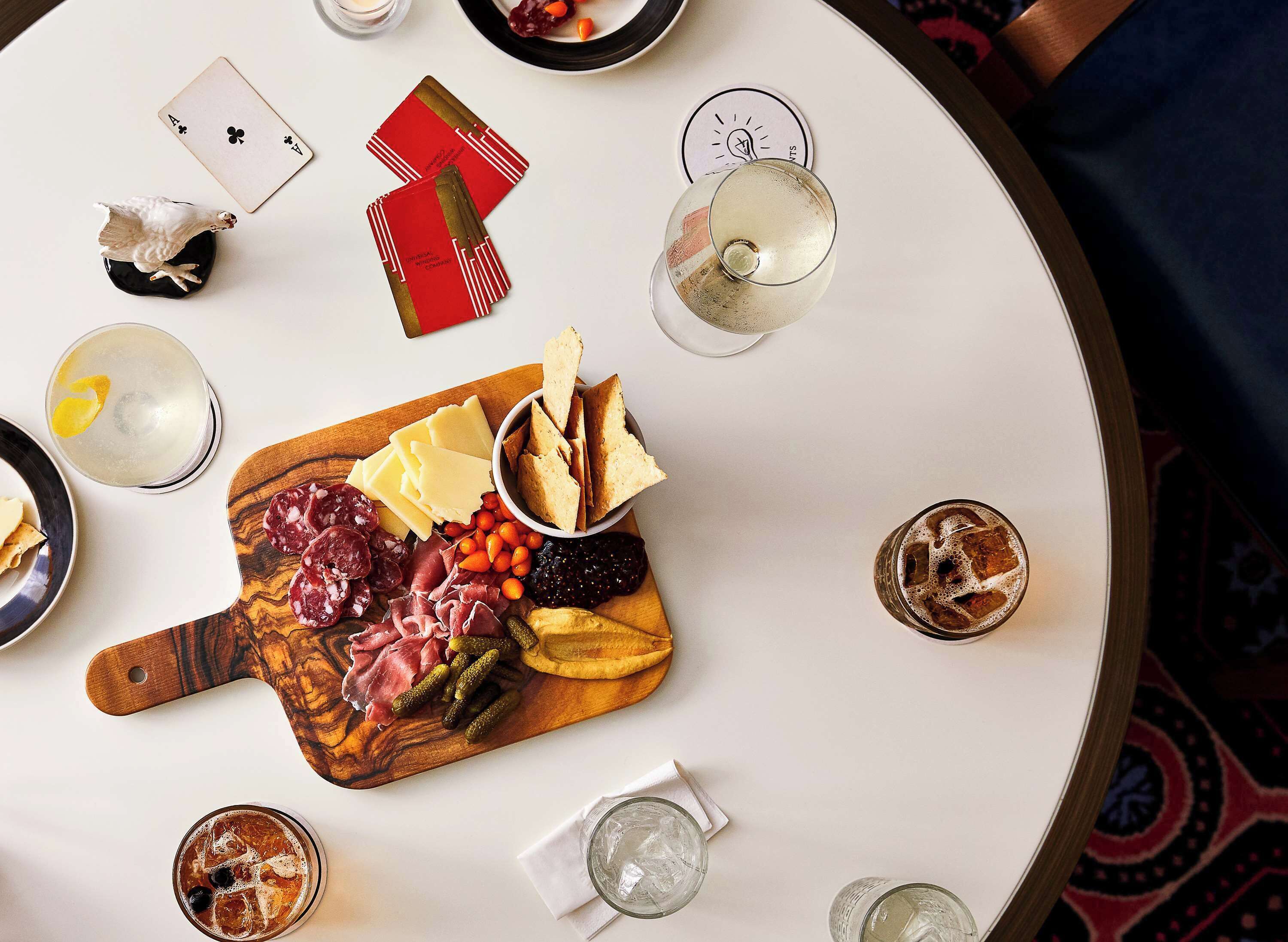 Une table ronde sur laquelle sont disposés divers aliments, notamment des fromages, des charcuteries et des biscuits secs, ainsi que plusieurs verres remplis de boissons.