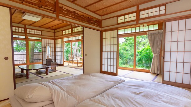 A traditional Japanese room with tatami flooring, a futon bed, sliding shoji doors, and a view of greenery outside.