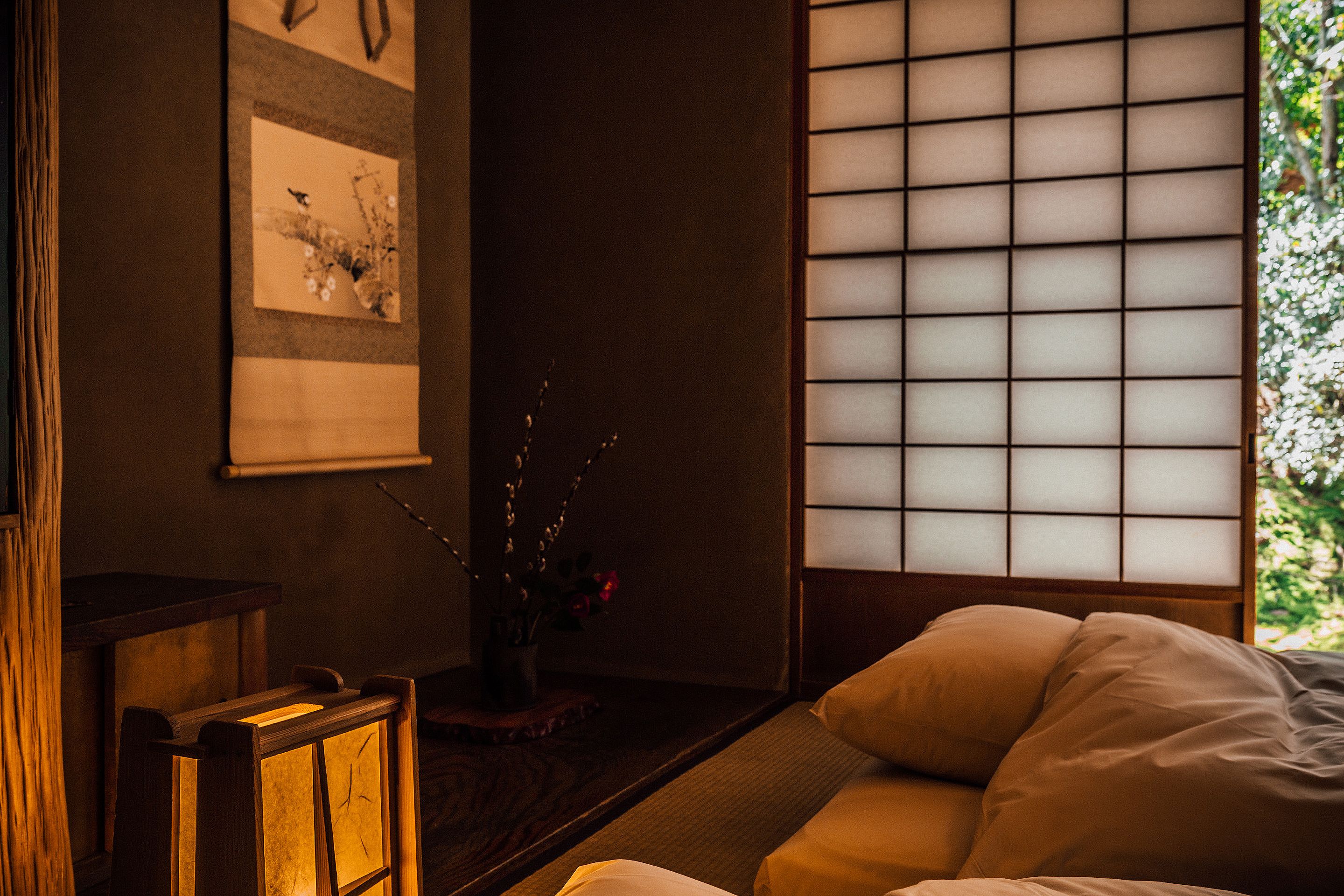 A traditional Japanese-style room with tatami flooring, a low wooden table, a shoji screen, and a bed with white bedding.