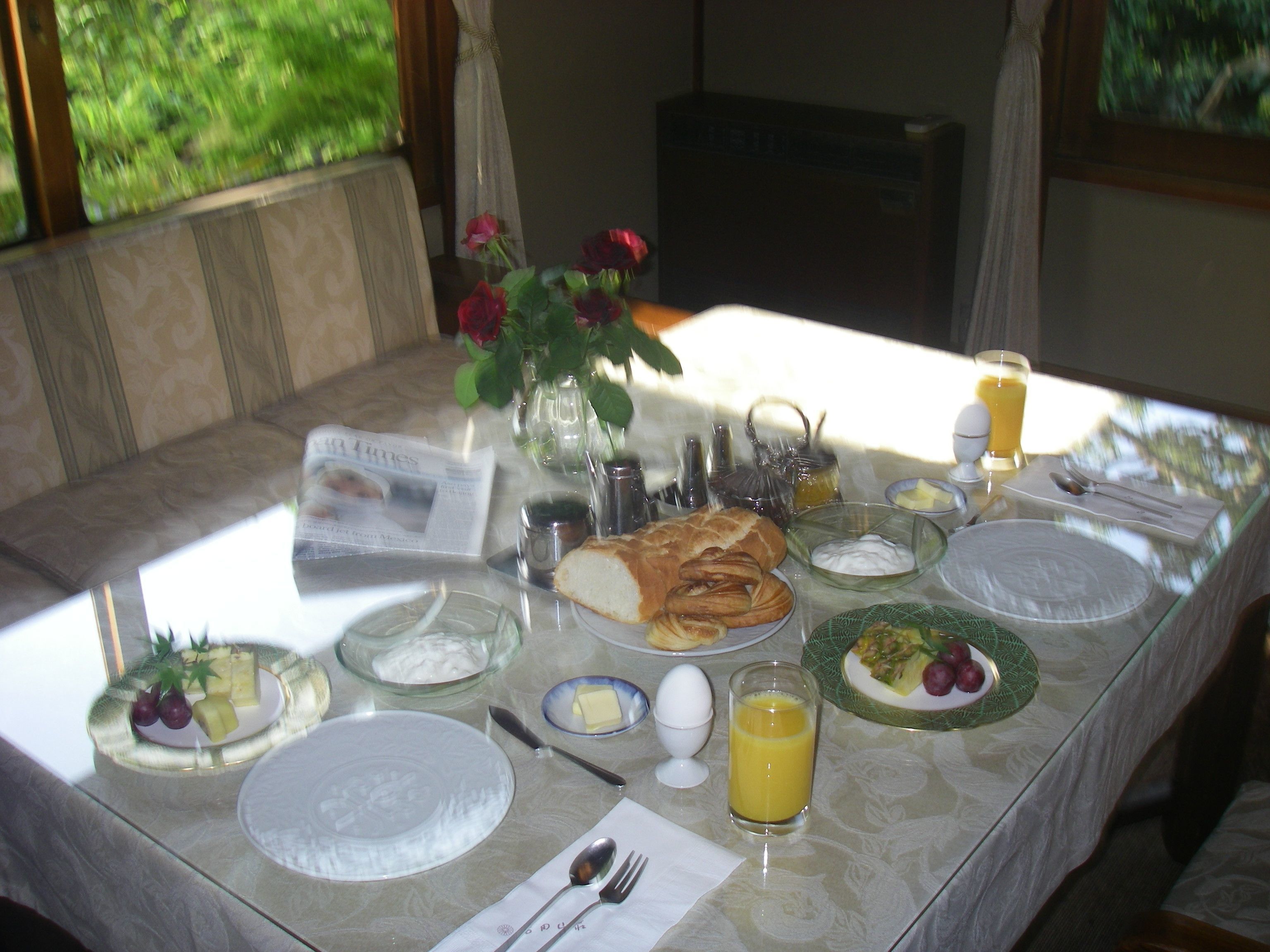 Mesa de café da manhã posta com pratos, pão, ovos, frutas e um copo de suco de laranja.
