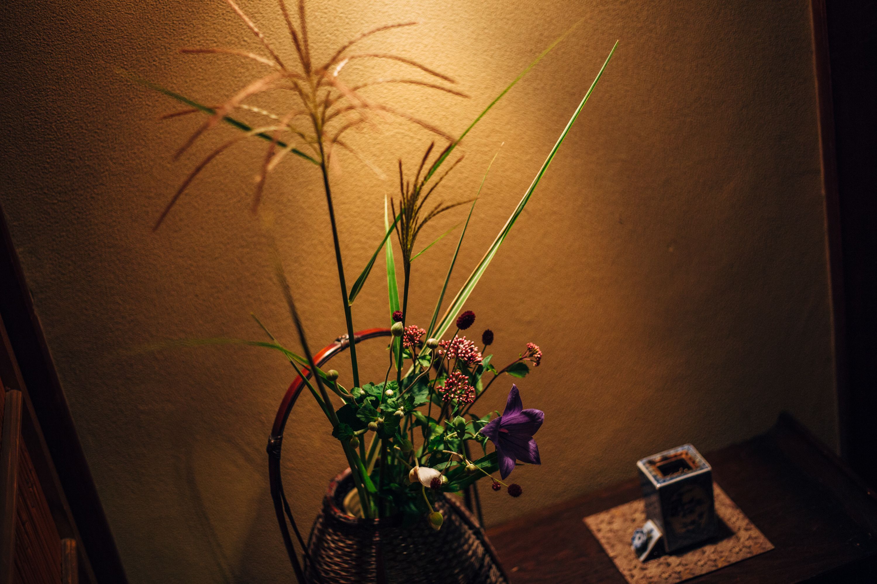 Un vase en osier, agrémenté d’une composition florale, est posé sur une table en bois devant un mur à texture.