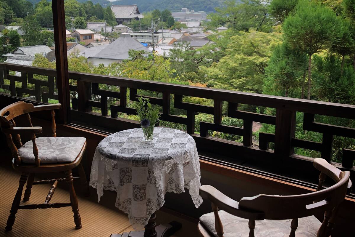 Ein traditioneller japanischer Raum mit Tatami-Böden, einem niedrigen Tisch mit weißen Spitzen, zwei Holzsesseln und Blick auf eine Stadt und Berge.