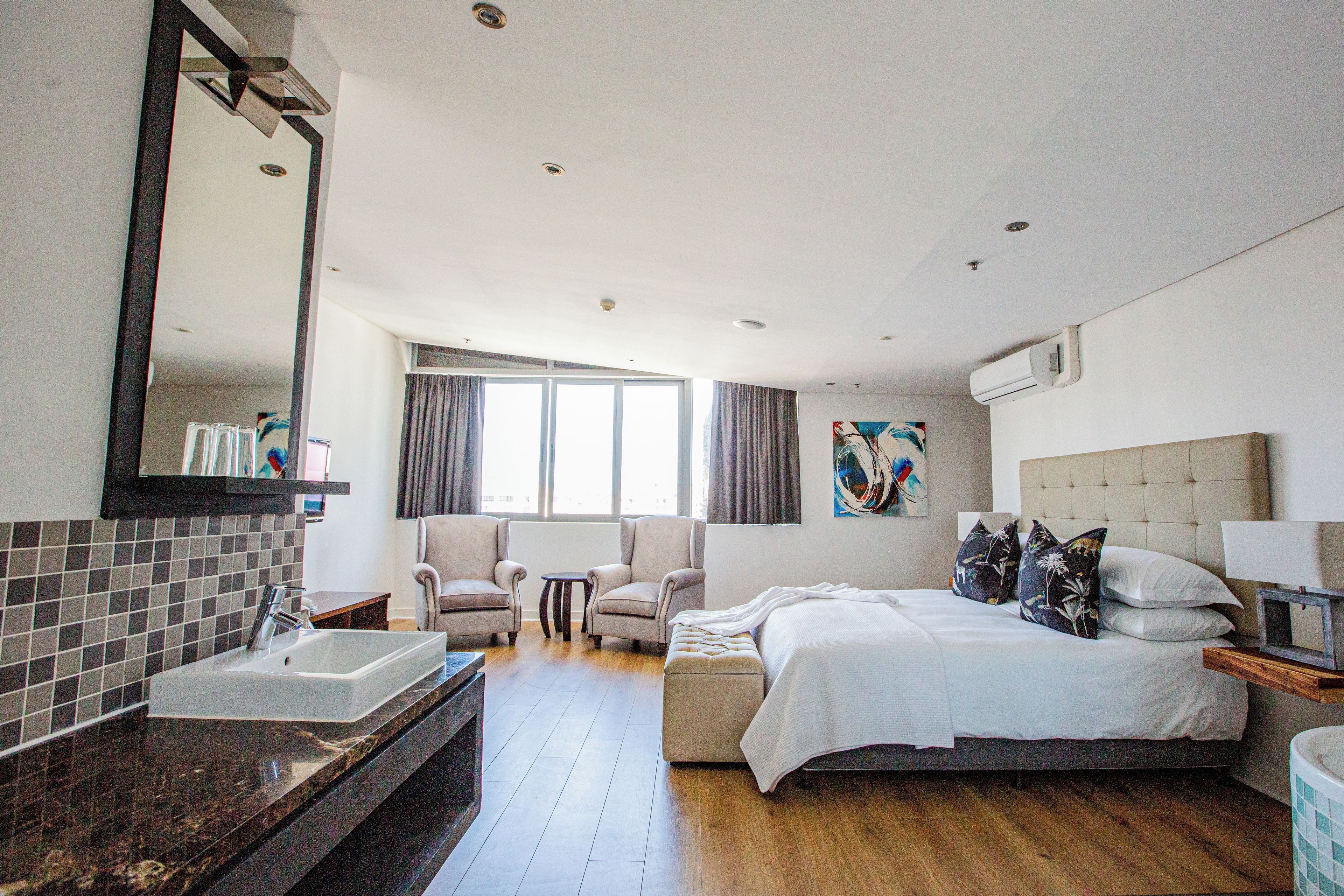 Una habitación de hotel con una cama grande, un área de descanso con dos sillones, un ventanal amplio, un baño con lavamanos y espejo, y un equipo de aire acondicionado.