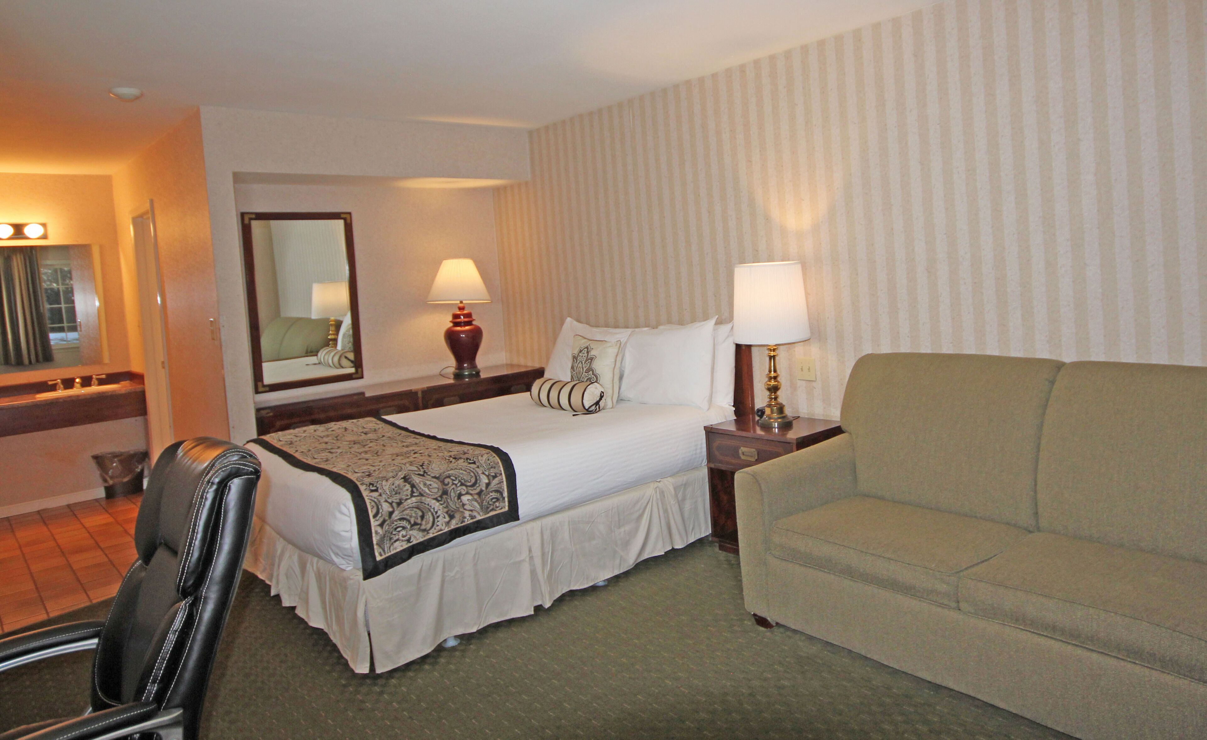 A hotel room with a bed, two bedside lamps, a mirror, a sofa, and a chair.