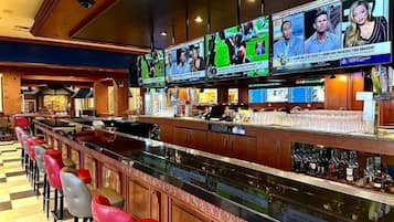 A bar area with multiple screens displaying sports content, a long counter with stools, and a variety of bottles on the shelves.