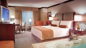 A hotel room with a large bed, a desk with a computer, a chair, a small table with a lamp, a window with curtains, and a bathtub.