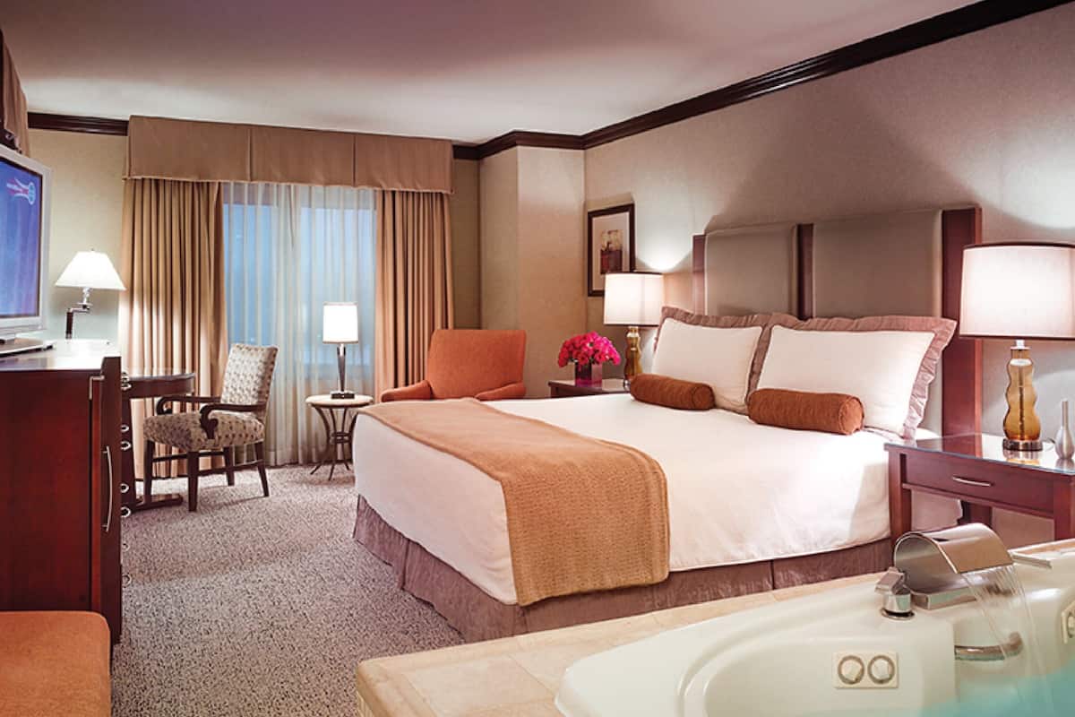 A hotel room with a large bed, a desk with a computer, a chair, a small table with a lamp, a window with curtains, and a bathtub.