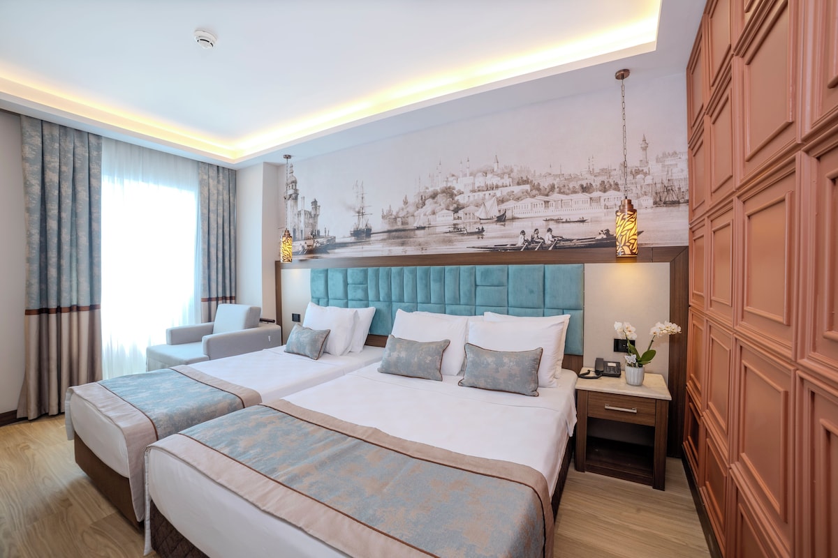 A hotel room with two beds, a large mural on the wall, a wooden wardrobe, and a nightstand with a lamp.